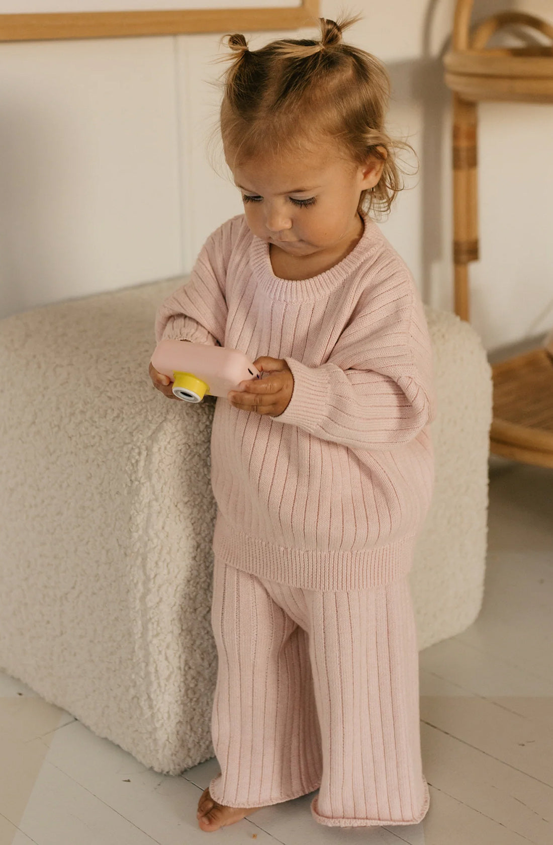 A toddler with light brown hair stands barefoot on a wooden floor. Dressed in a cozy Junee Knit Sweater Macaron with matching pants, both from GOLDEN CHILDREN and made of 100% cotton knit, the child intently examines a small, yellow and pink toy camera. The background includes a plush, textured ottoman and a wicker chair.