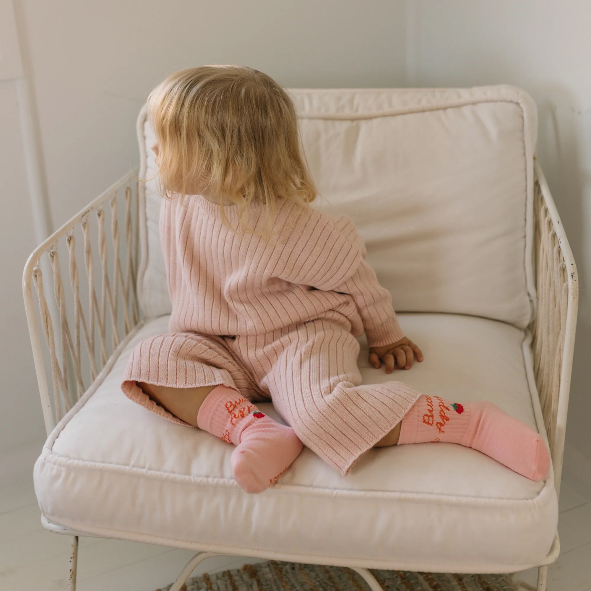 A blonde toddler, dressed in a ribbed, light pink long-sleeve outfit and pink socks, sits on a white cushioned chair facing away and looking to the side. The 100% cotton knit ensemble from GOLDEN CHILDREN ensures comfort. The room's white walls and light décor create a soft and peaceful ambiance reminiscent of an ad for the Junee Knit Sweater Macaron.