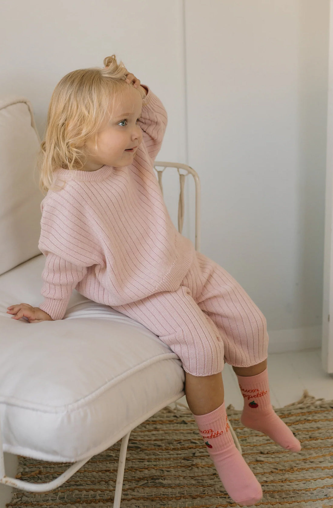 A young child with blonde hair sits on the edge of a white couch, wearing a relaxed fit Junee Knit Sweater Macaron by GOLDEN CHILDREN, made from 100% cotton knit. The pink ensemble includes the sweater and matching pants, complemented by pink socks adorned with small designs. The child gazes off to the side with one hand slightly raised to their head, appearing relaxed.