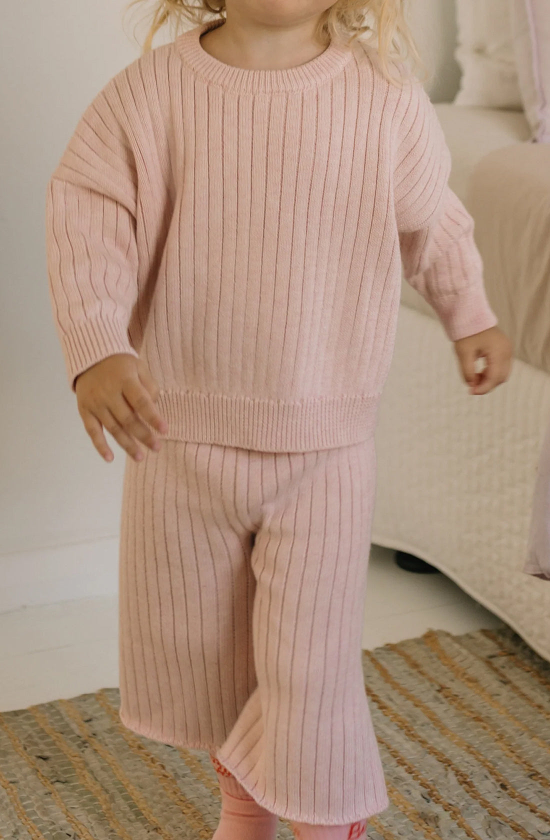 A young child with light blond hair is walking indoors, wearing a matching pink Junee Knit Sweater Macaron and pants from GOLDEN CHILDREN. The outfit is made from 100% cotton knit for a relaxed fit. The child is moving towards the right side of the image, with a bed featuring white bedding and a beige woven rug in the background.