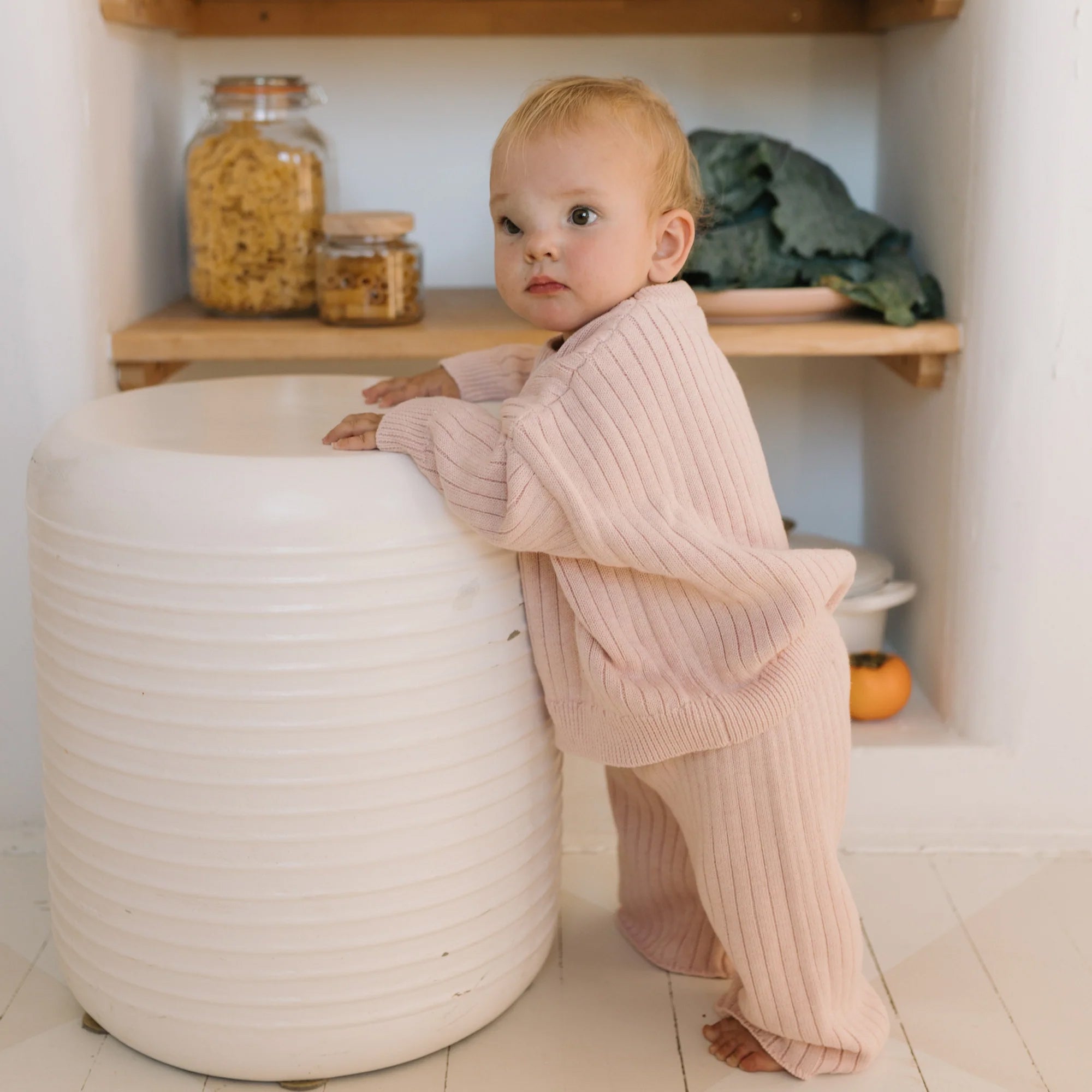 A young child with short blonde hair is standing and holding onto a white ribbed object in a cozy kitchen setting. The child is dressed in the matching light pink 100% cotton Junee Knit Sweater Macaron and pants from GOLDEN CHILDREN, featuring a relaxed fit. Shelves in the background contain jars of pasta, cookies, and leafy vegetables.