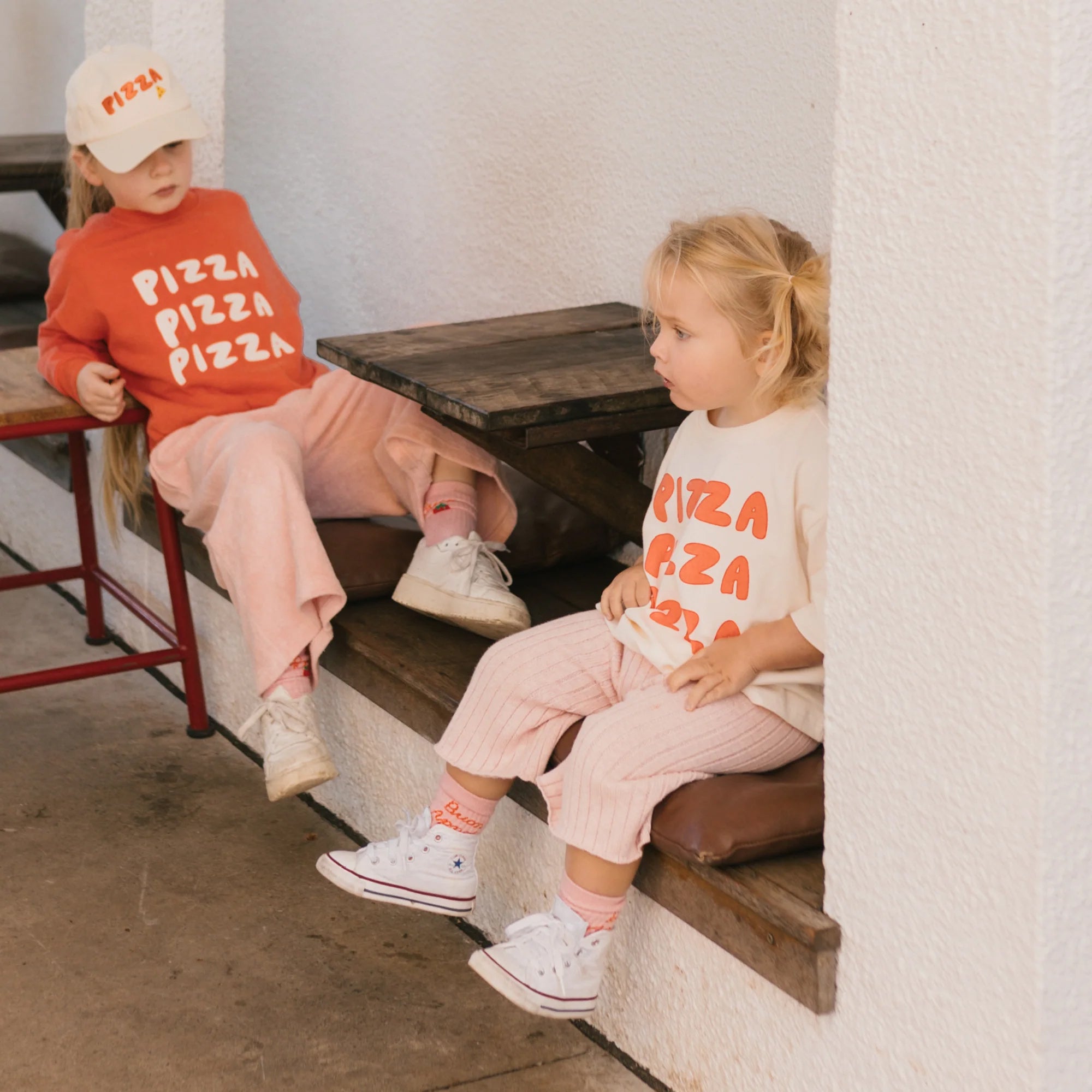 Two young children sit on wooden benches by a white wall. Both are wearing "PIZZA PIZZA PIZZA" sweatshirts, one in an oversized fit in red and the other in white, paired with pink GOLDEN CHILDREN Junee Knit Wide Leg Pants Macaron. They both have blonde hair and are looking off into the distance. One child wears a matching cap.