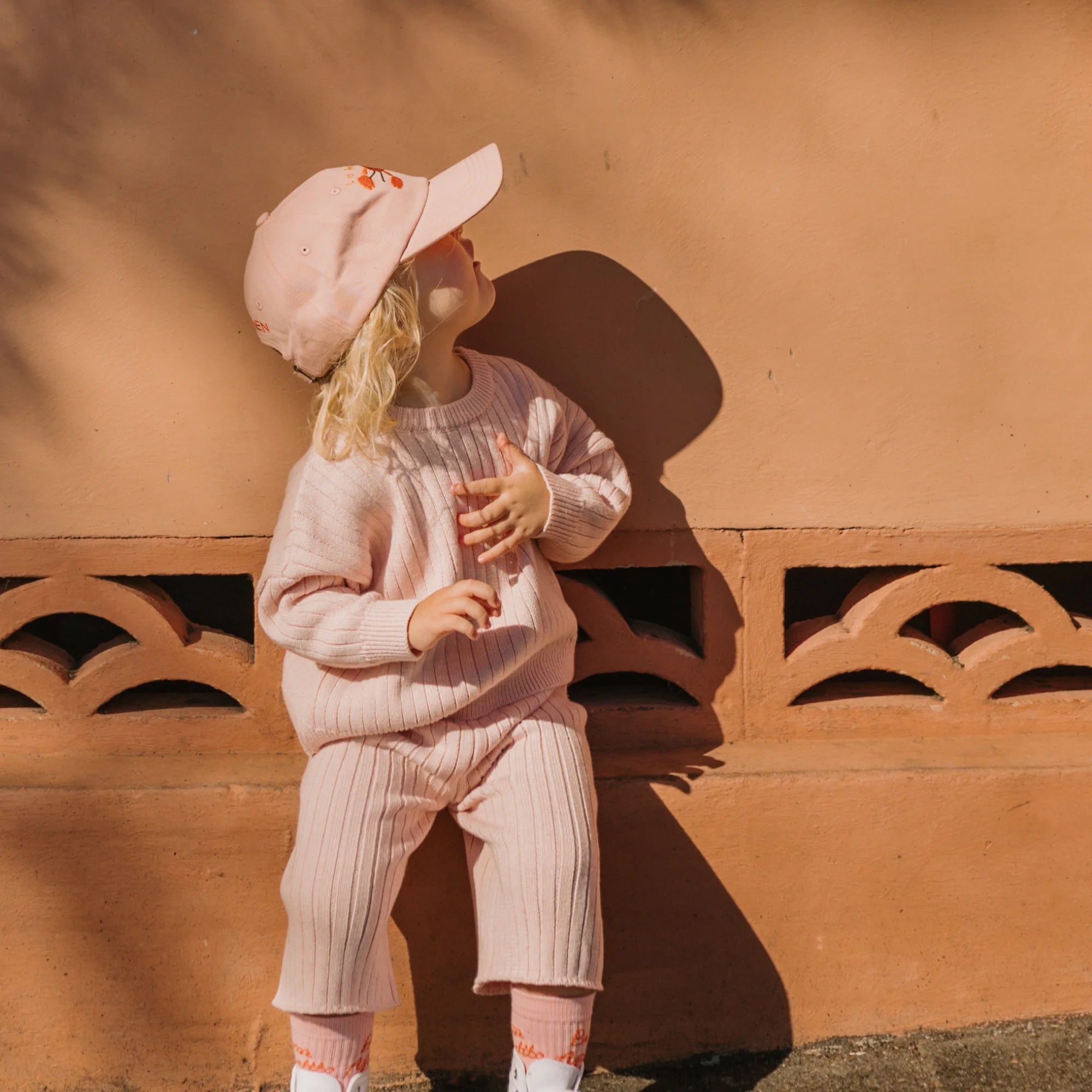 A young child with blonde hair is dressed in a 100% cotton knit, pink Junee Knit Sweater Macaron by GOLDEN CHILDREN, along with matching pants and a pink baseball cap. The relaxed fit outfit complements the child as they look to the side while leaning against a decorative terracotta-colored wall, partially shaded by nearby trees.