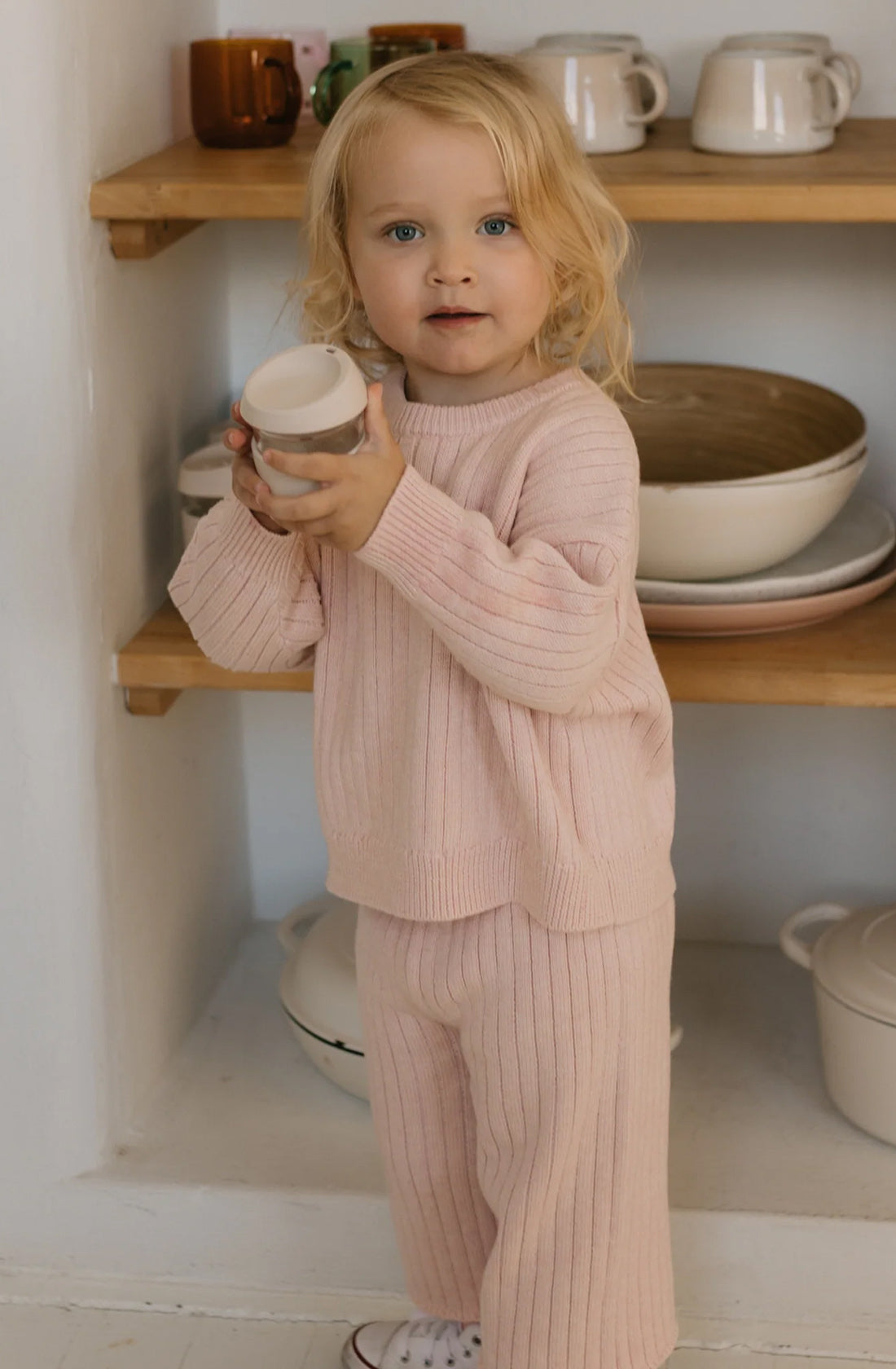 A young child with light blonde hair stands in a kitchen, holding a small white cup. They are wearing a cozy, pink Junee Knit Sweater Macaron and matching pants from GOLDEN CHILDREN. This relaxed-fit attire is made of 100% cotton knit. Shelves in the background hold various cups, bowls, and a teapot. The child looks directly at the camera.