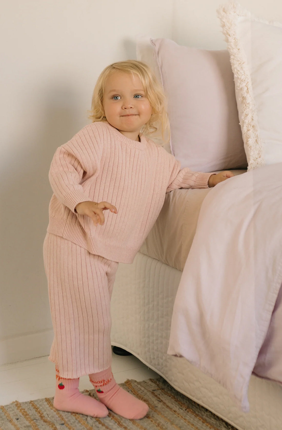 A young child with blonde hair stands beside a bed, wearing a cozy, light pink Junee Knit Sweater Macaron from GOLDEN CHILDREN and matching pants made from 100% cotton knit. The relaxed fit ensemble includes pink socks with red accents, and the child has a cheerful expression on their face in the softly neutral-toned room.