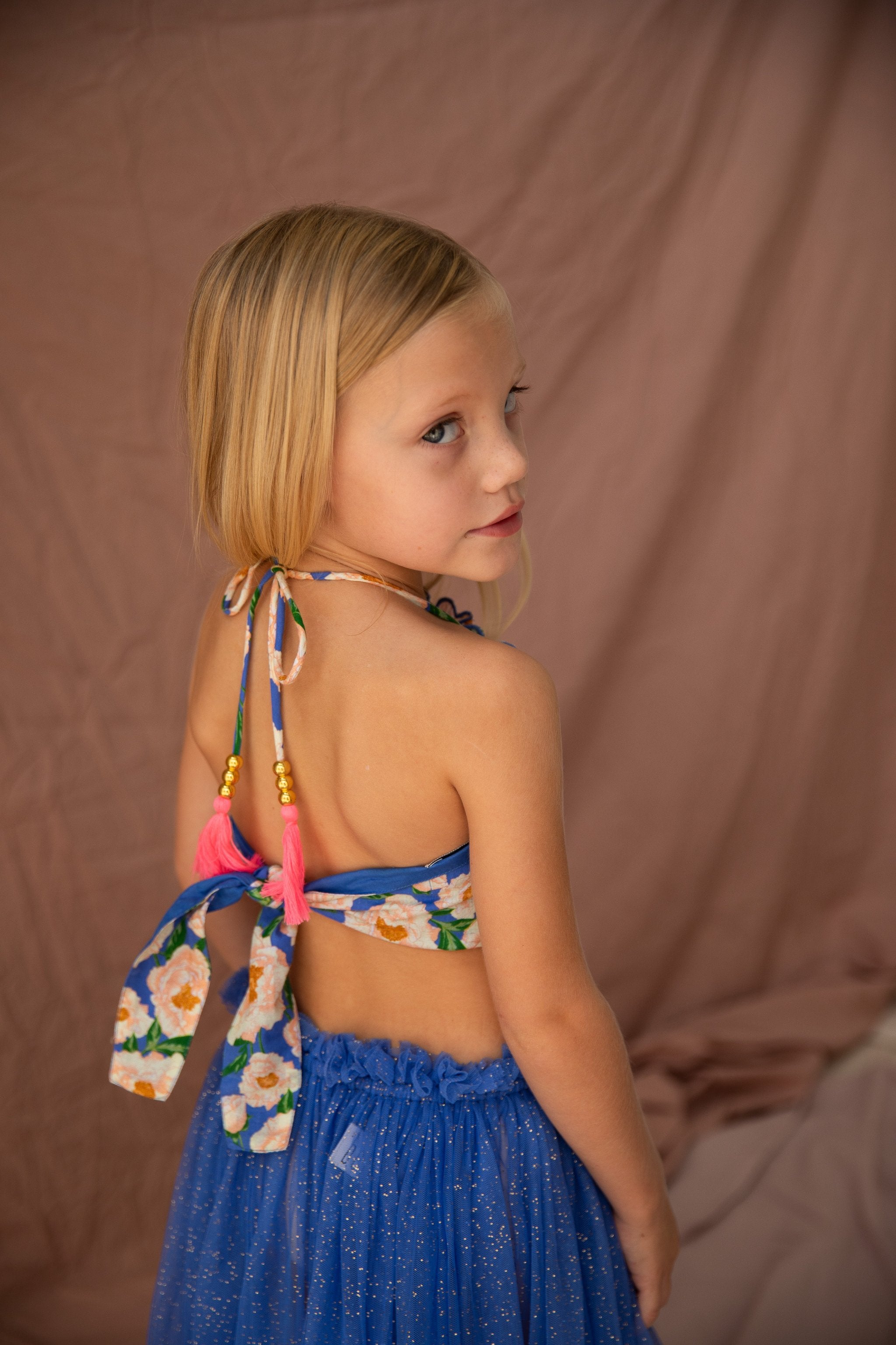 A young girl with blonde hair, wearing the BELLA + LACE Jess Top Soirée Bouquet/Bondi Blue and a sparkling blue skirt with fluttery shoulders, stands in front of a beige fabric backdrop. She is looking over her shoulder towards the camera.