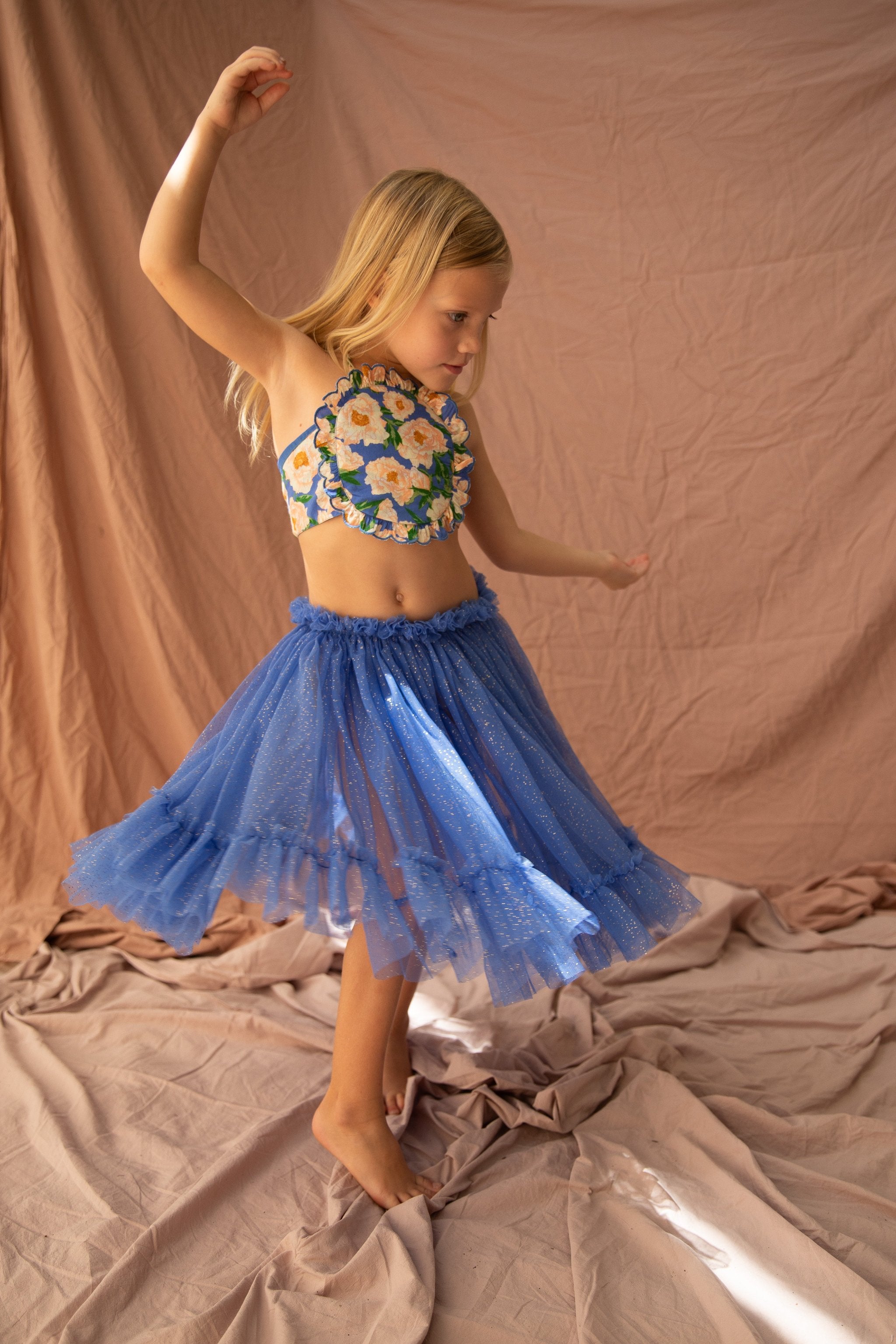 A young girl with blonde hair dances barefoot on a beige fabric backdrop. She is wearing the Jess Top Soirée Bouquet/Bondi Blue by BELLA + LACE, featuring a flower pattern, paired with a sparkling blue tulle skirt adorned with delicate scalloped embroidery. One arm is raised above her head while the other is outstretched to her side as she twirls.