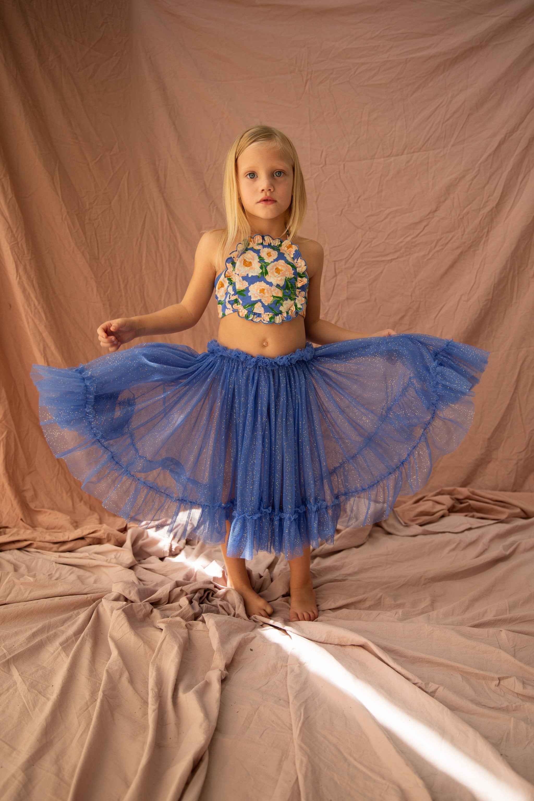 A young child stands barefoot on a draped fabric backdrop, wearing the BELLA + LACE Jess Top Soirée Bouquet/Bondi Blue, a floral-patterned crop top with fluttery shoulders, and a blue tulle skirt. They are holding the skirt out in both hands, creating a fan-like shape. The backdrop is a soft blush color.