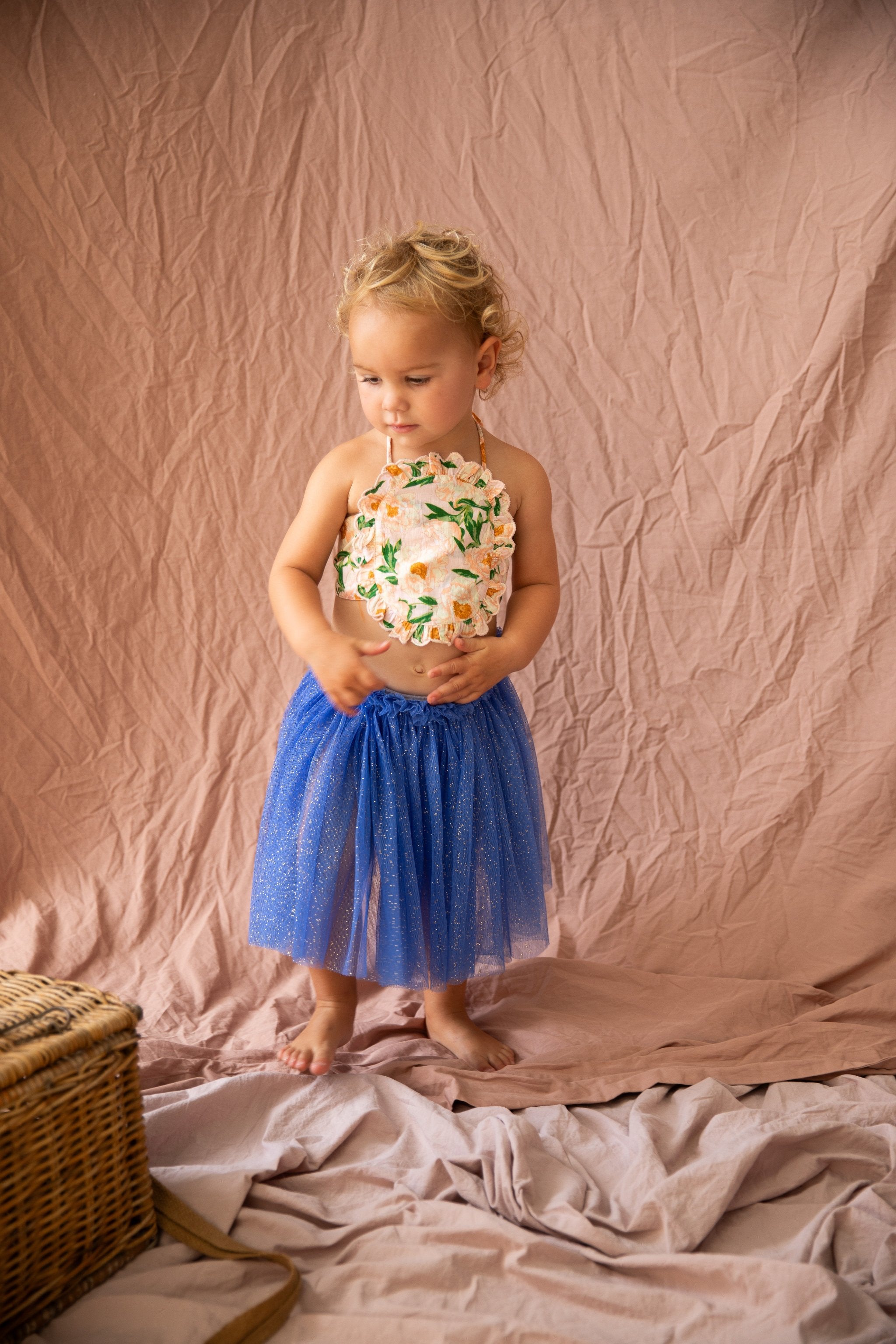 A toddler with curly blond hair stands barefoot on a crumpled pink sheet. She is wearing a floral halter top and the Classic Tutu Bondi Blue from BELLA + LACE, an extra soft tulle skirt decorated with glitter. To her left sits an open wicker basket. She looks down, appearing lost in thought, embodying the charm of BELLA + LACE attire.