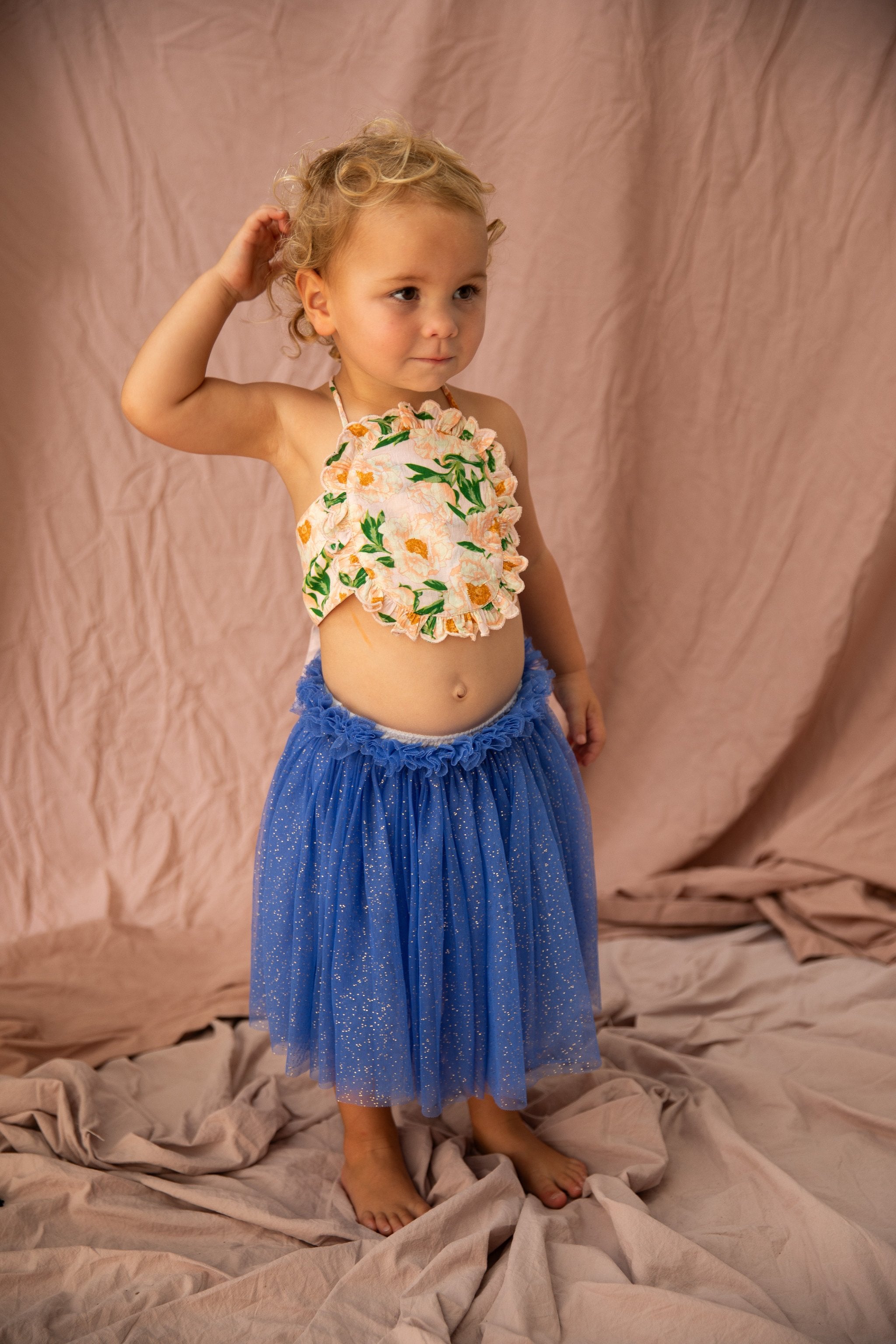 A toddler with blonde curly hair stands barefoot on a crumpled cloth, wearing a floral top and a BELLA + LACE Classic Tutu in Bondi Blue. The background is a light pink fabric. The child, clad in an extra soft tulle skirt, is looking to the side with one arm raised to their head.