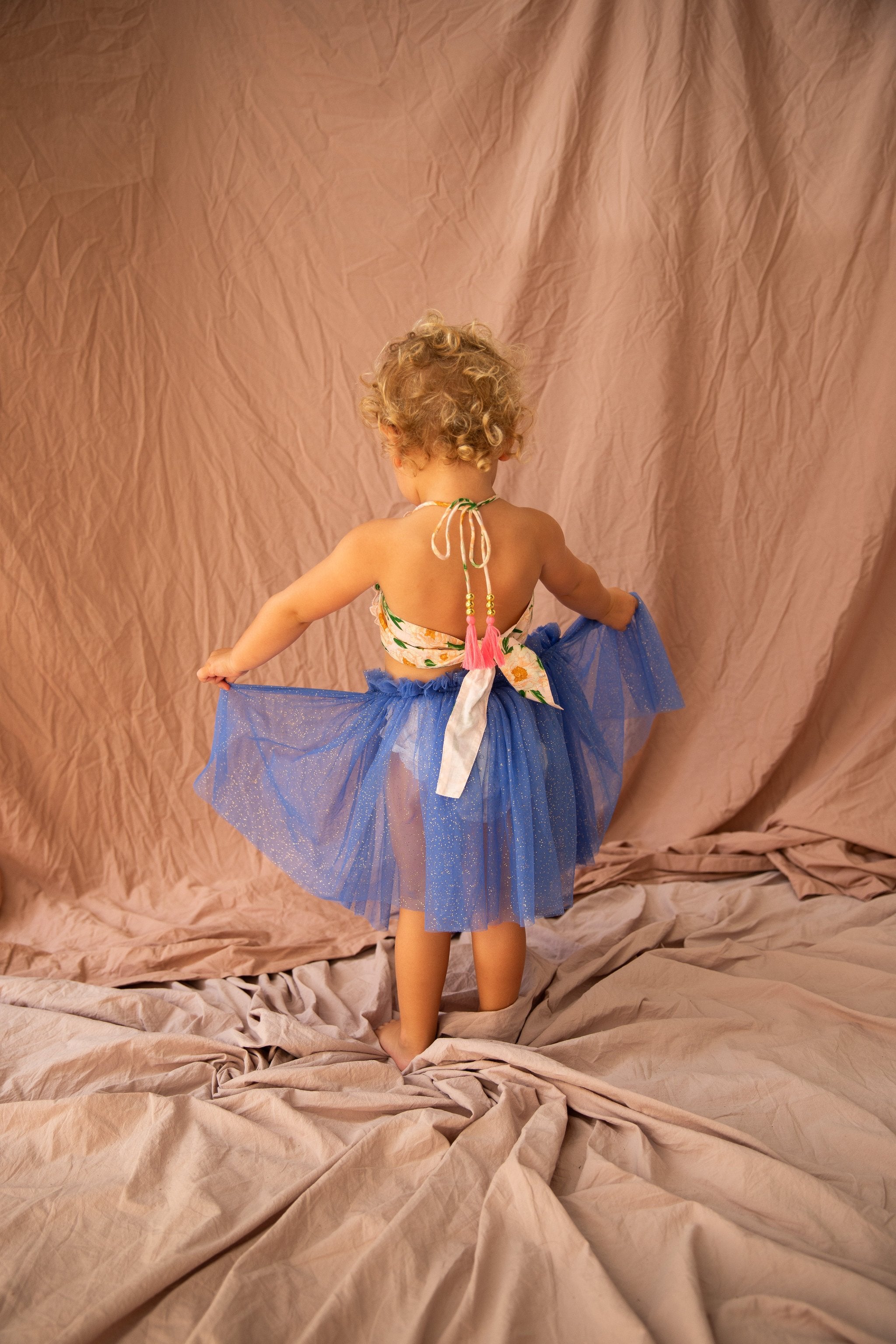 A toddler with curly blonde hair stands facing away, wearing the Classic Tutu Bondi Blue by BELLA + LACE. The blue sparkly tutu dress, featuring a floral bodice, is made of extra soft tulle. The background consists of softly draped wrinkled pink fabric. The child holds the sides of the tutu, slightly lifting it.