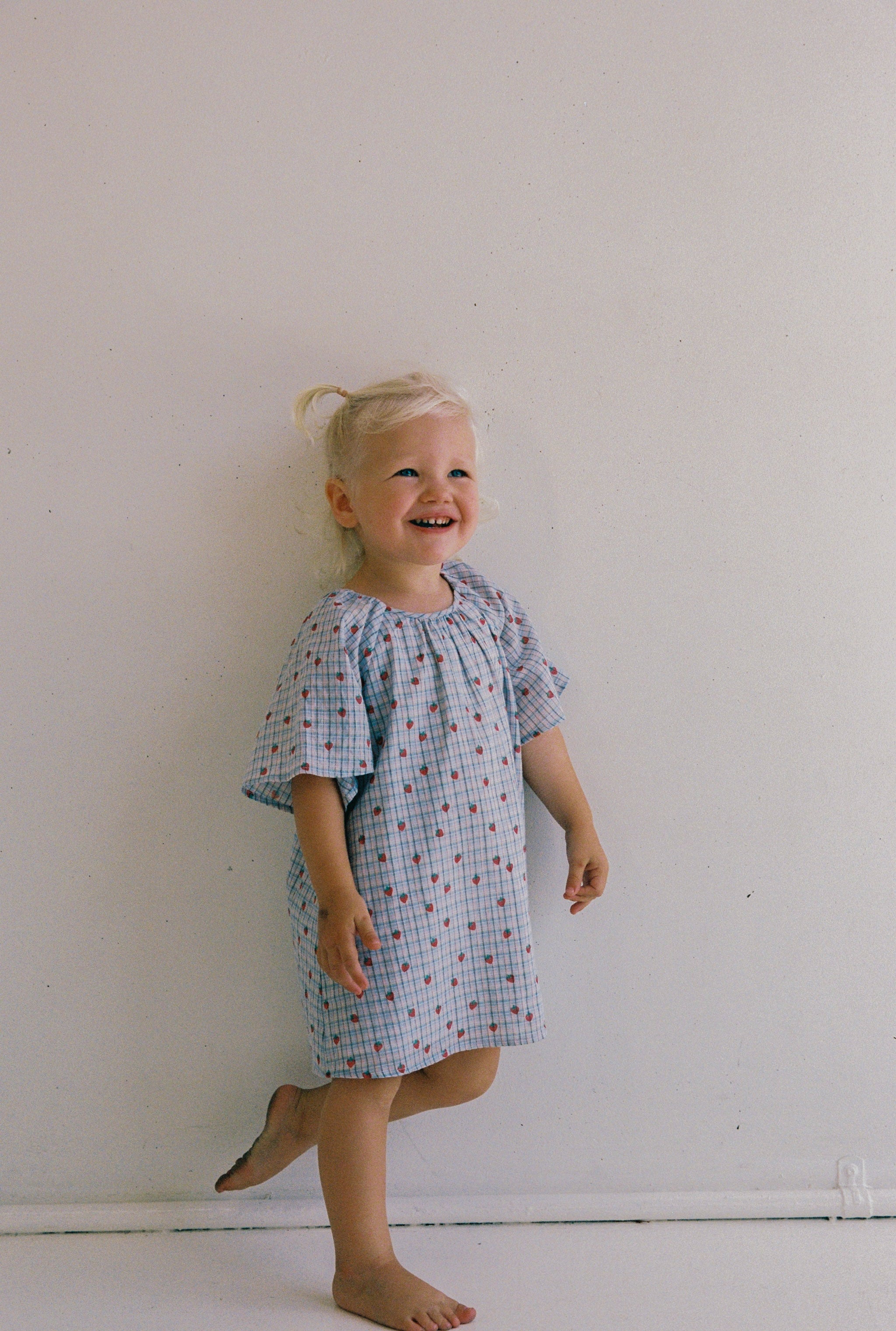 A young child with light blonde hair in pigtails, smiling and looking upward, wears a JUNI JNR Penny Dress Strawberry Check (Size 4) and stands barefoot against a plain white wall.