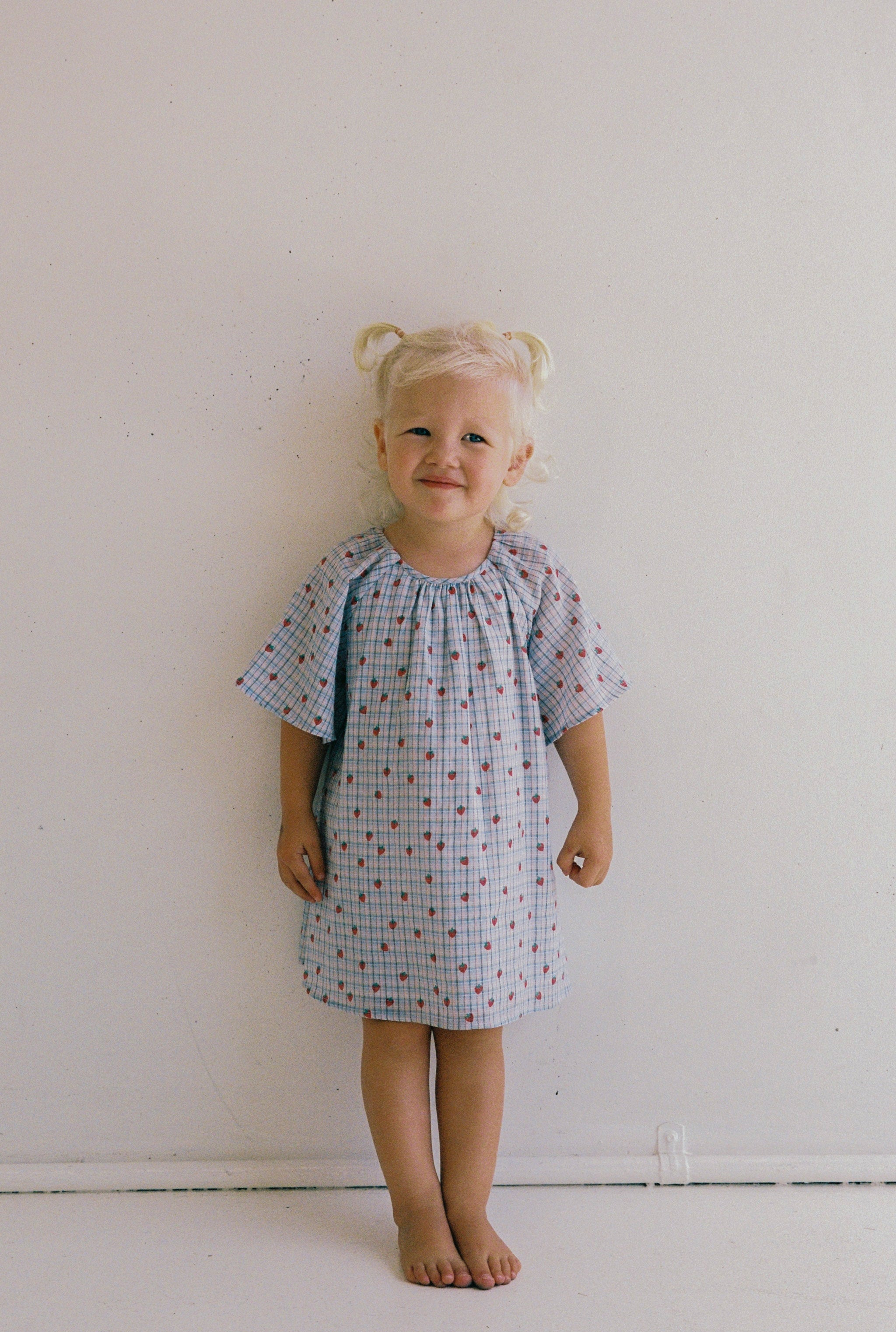 A young child with blonde hair in pigtails stands against a white wall, wearing the ethically made Penny Dress Strawberry Check - Size 4 from JUNI JNR. The light blue dress, adorned with red and blue geometric patterns, features a gathered neckline. They're barefoot and smiling brightly.