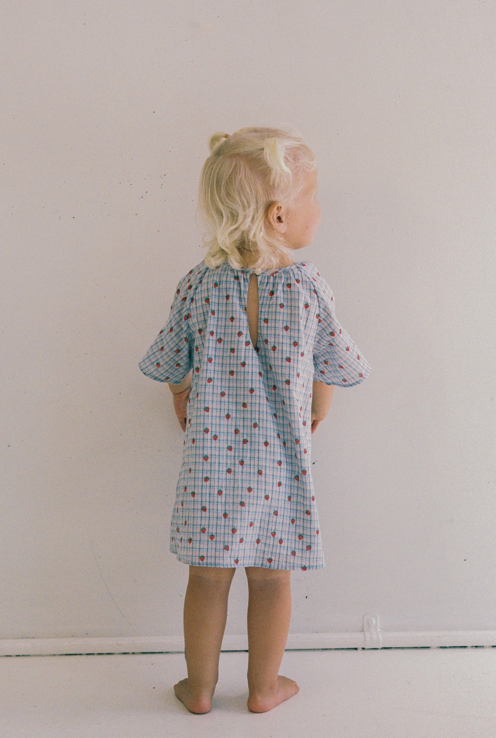 A small blonde child stands barefoot against a white wall, facing away. The child is wearing the Penny Dress Strawberry Check by JUNI JNR, an ethically made light blue dress with red dots and floral patterns, styled with two short ponytails.