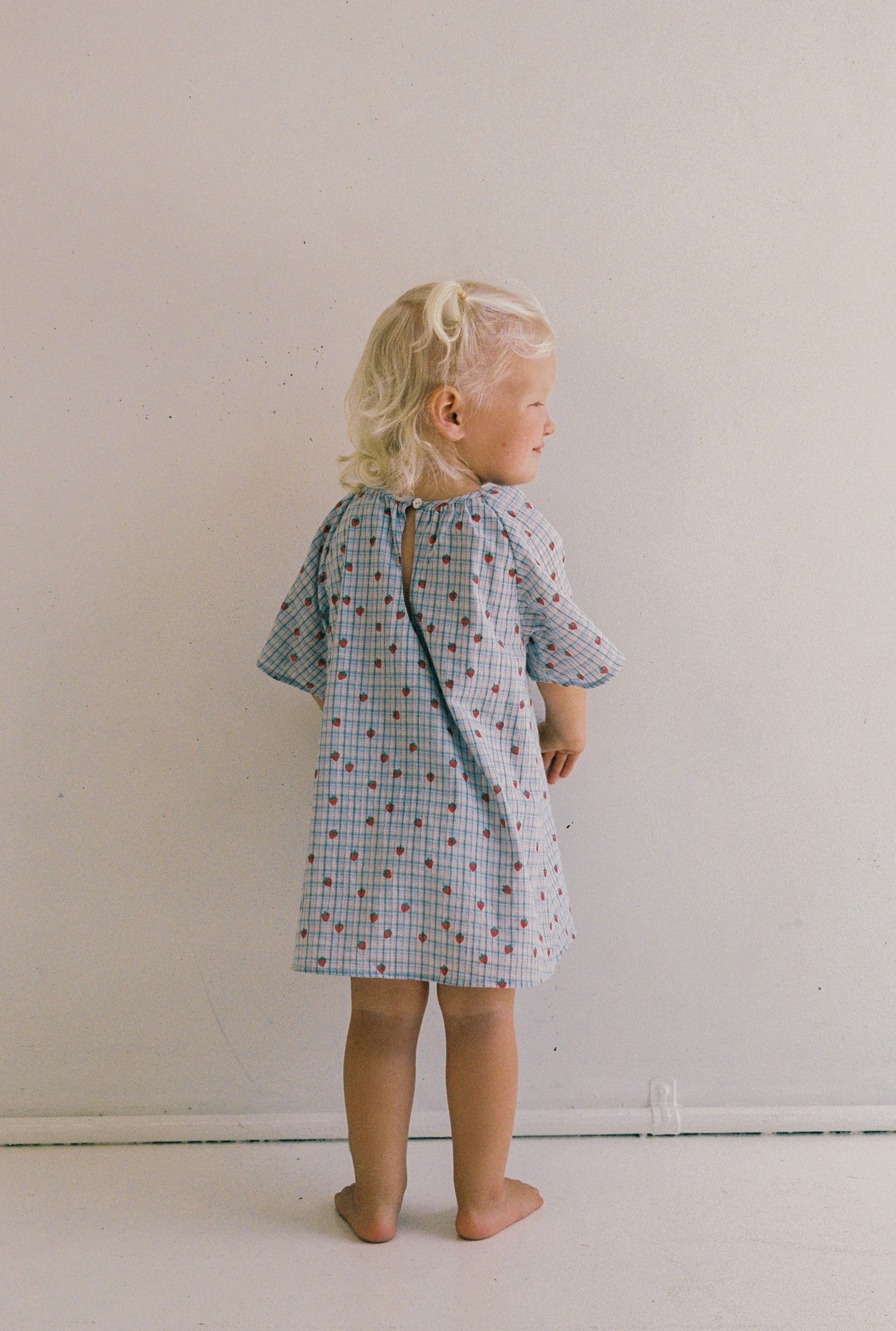 A young blonde child stands barefoot against a white wall, wearing the JUNI JNR Penny Dress Strawberry Check in Size 4. The short-sleeved dress features small red patterns and a charming gathered neckline. The child faces away, slightly glancing sideways, embodying innocence and style.