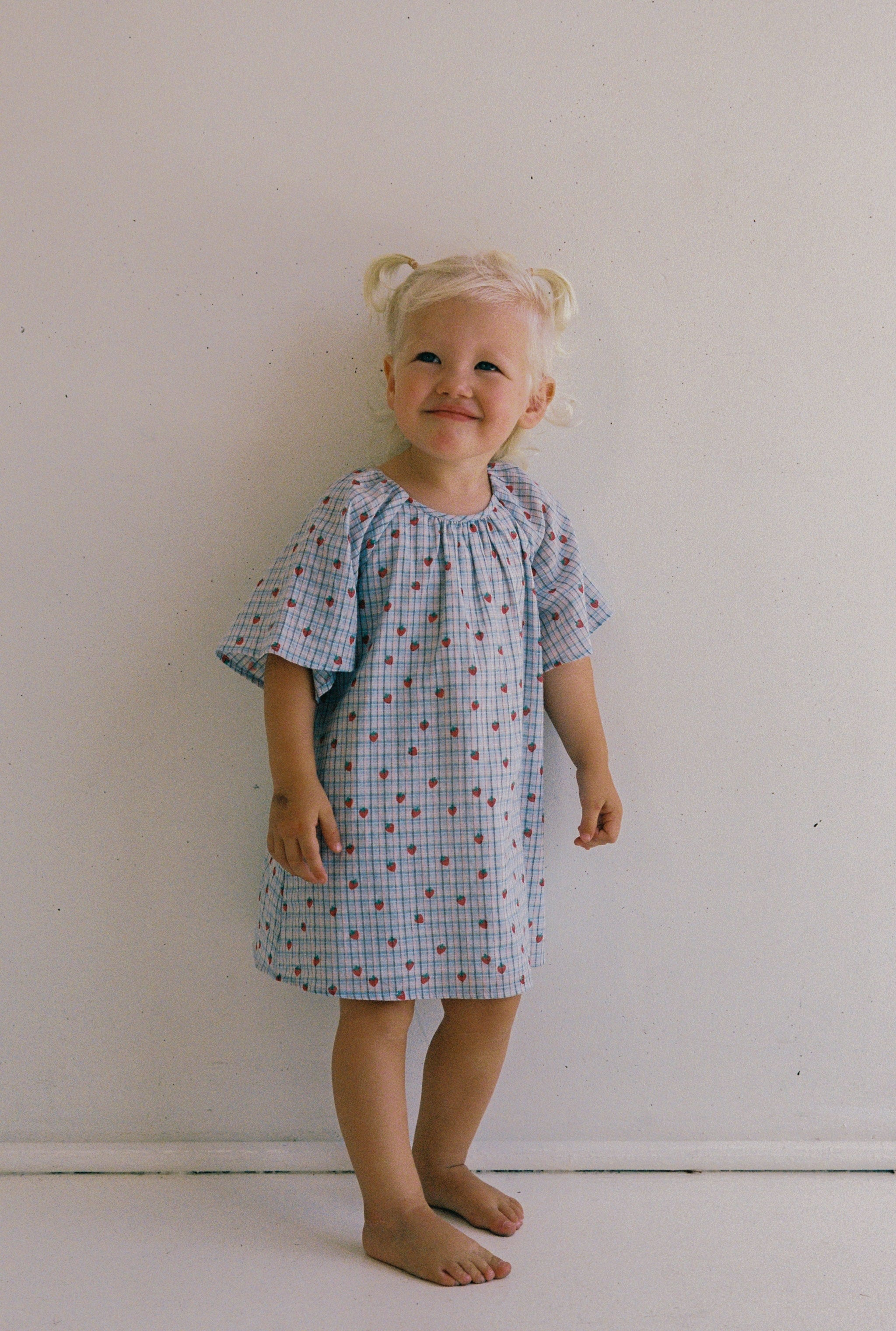 A young child with blonde hair in small buns stands barefoot against a plain background, wearing the ethically made JUNI JNR Penny Dress Strawberry Check in size 4. The light-colored dress features small red dots and a gathered neckline as the child smiles slightly and looks upward.