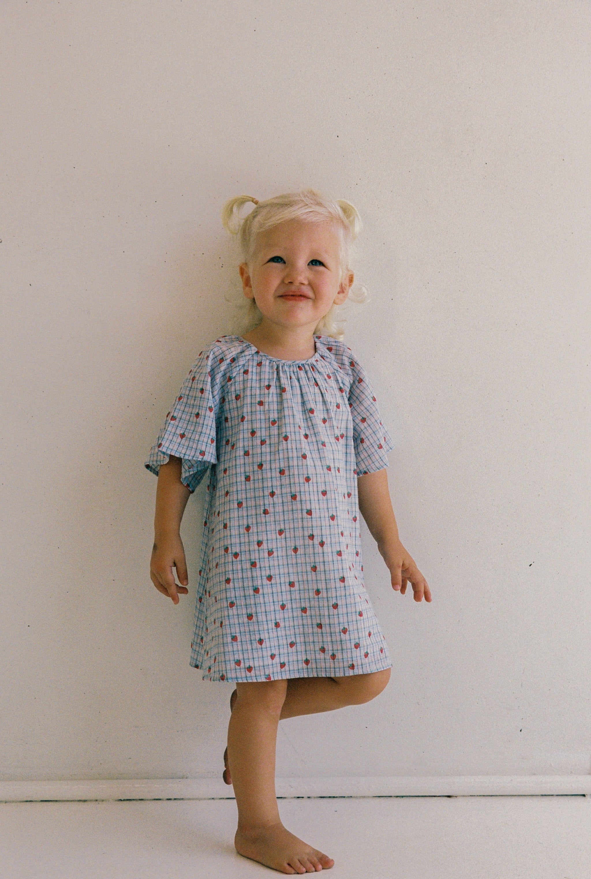 A young child with blonde pigtails smiles joyfully while barefoot against a plain white wall, wearing the Penny Dress Strawberry Check by JUNI JNR in size 4.