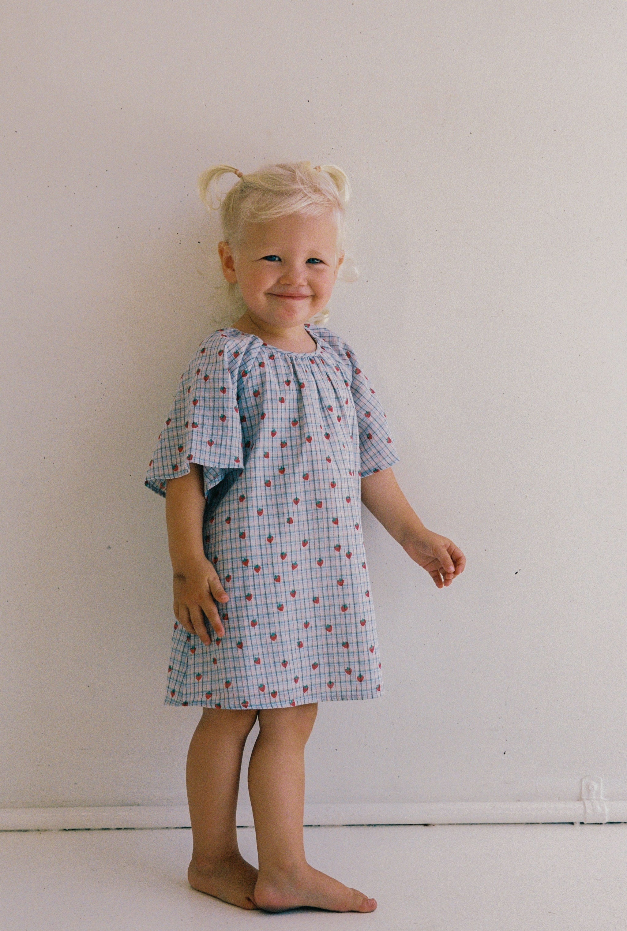 A young child with blonde hair in pigtails stands barefoot against a plain backdrop, wearing the Penny Dress Strawberry Check by JUNI JNR. The blue and white yarn dye check fabric features red patterns and a gathered neckline. They smile slightly while facing the camera.