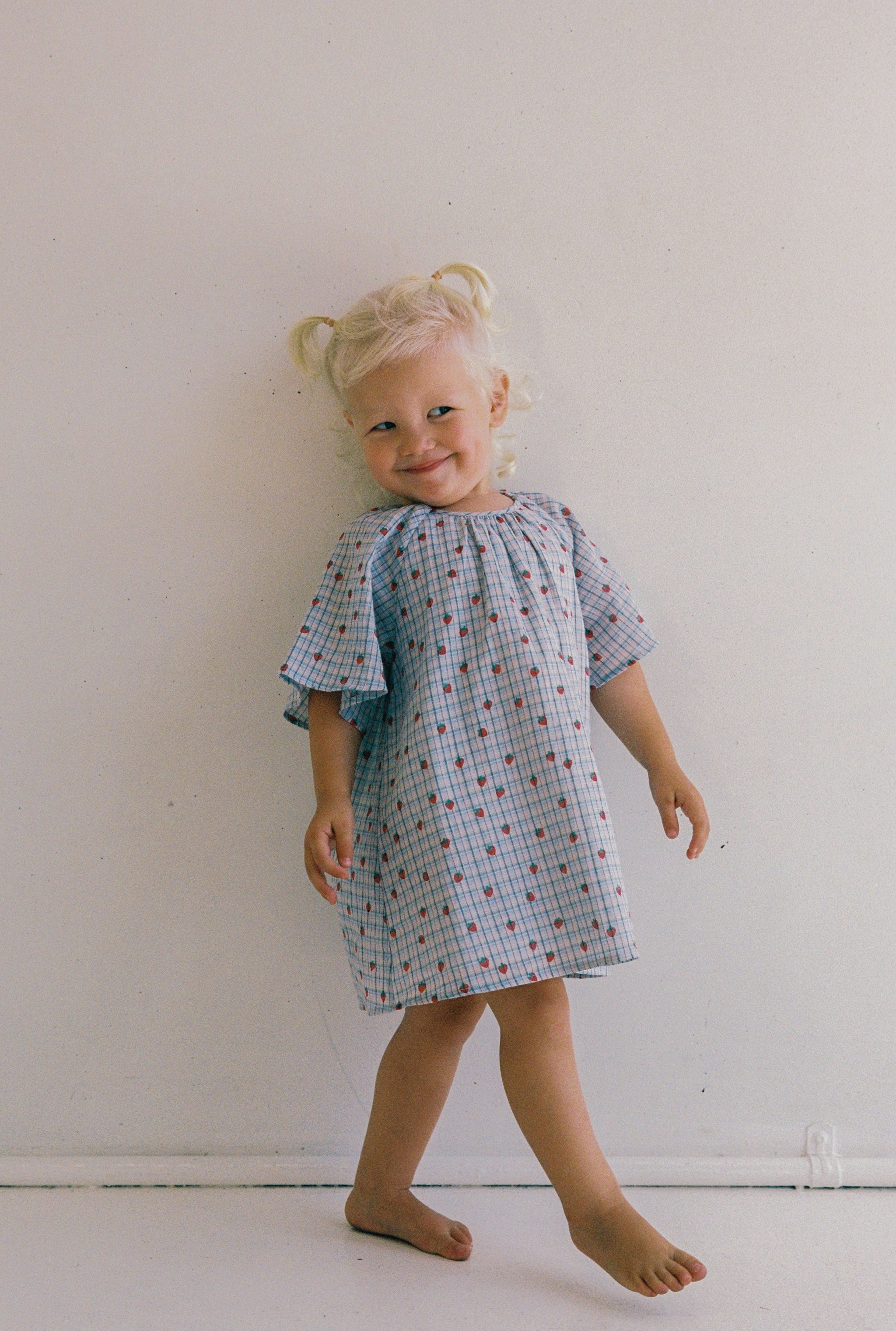 A joyful blonde girl stands barefoot before a white wall, dressed in the JUNI JNR Penny Dress Strawberry Check. The blue and white checkered yarn dye check dress has small red patterns and an ethical gathered neckline. She sports two pigtails.