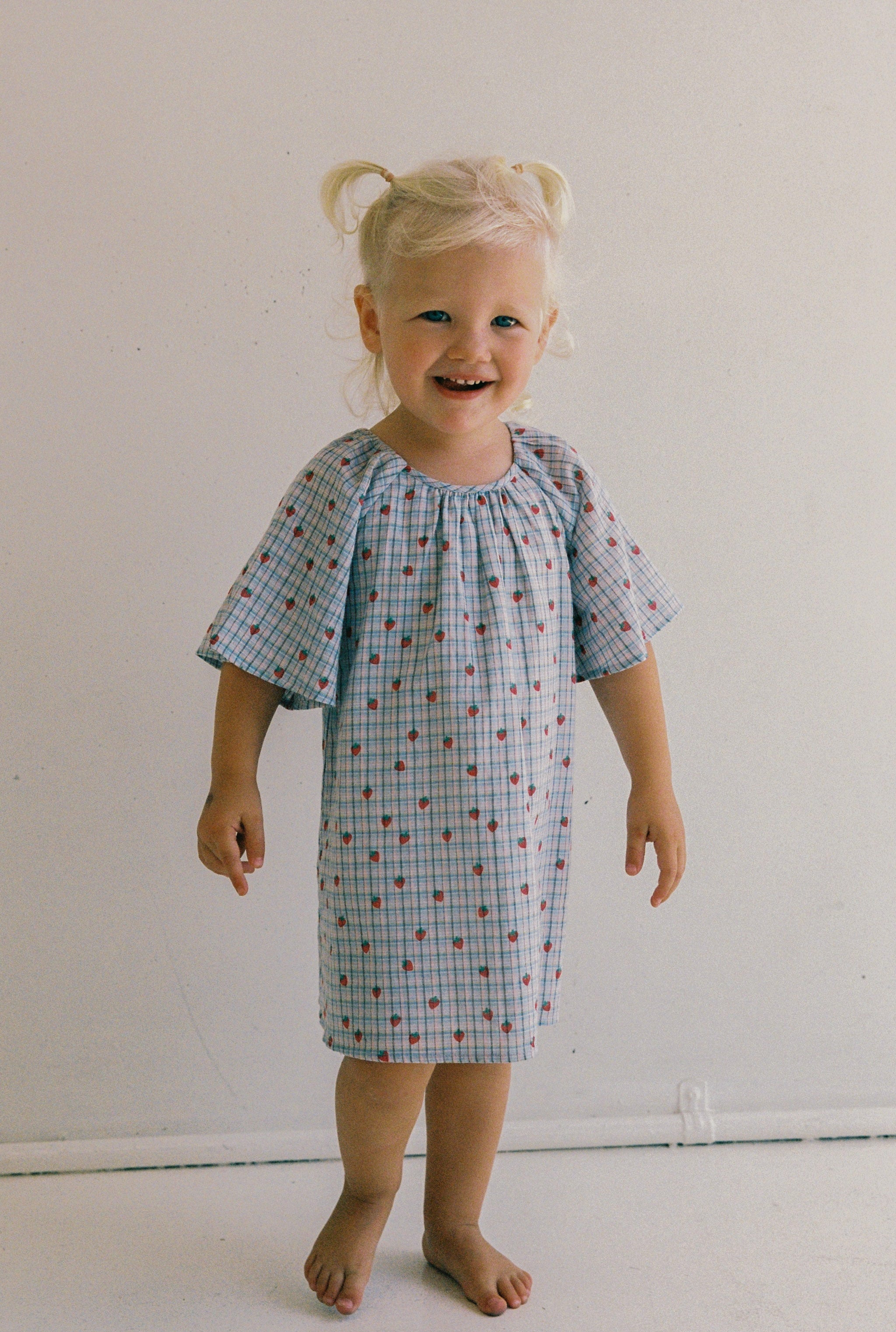 A smiling toddler with blonde pigtails stands barefoot on a plain background, wearing the JUNI JNR Penny Dress Strawberry Check in size 4. The dress is ethically made with a blue and white checkered pattern, red dots, and a gathered neckline.