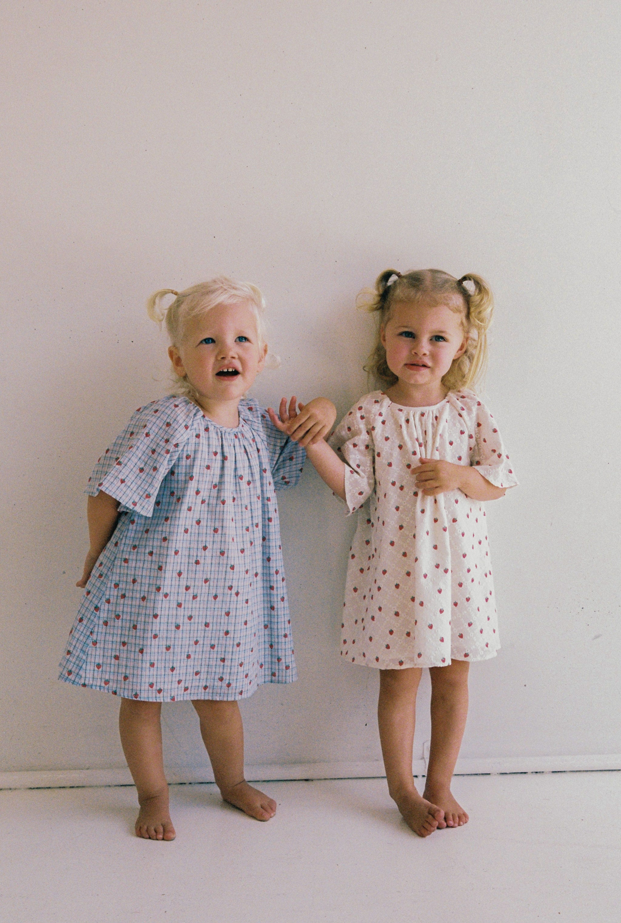 Two young children stand barefoot against a white wall, each in JUNI JNR dresses with gathered necklines—one in the Penny Dress Strawberry Check (Size 4) and one blue with red dots. Both have light-colored hair styled in pigtails.