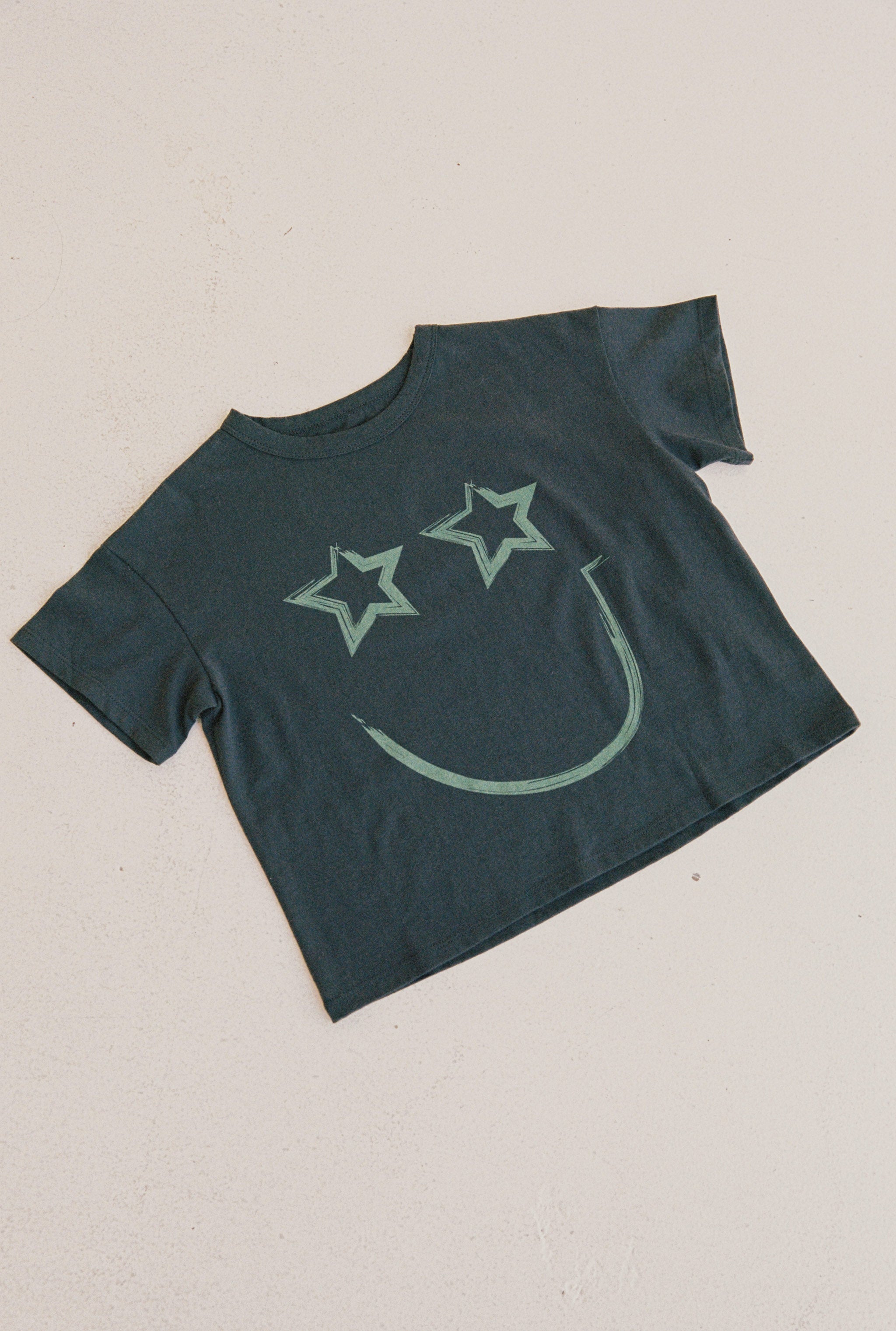 Presenting the Stellar Tee by JUNI JNR, a black T-shirt made from GOTS certified organic cotton. It showcases a smiley face with star-shaped eyes and a curved line mouth in faded green. This ethically produced piece is displayed flat on a white background.