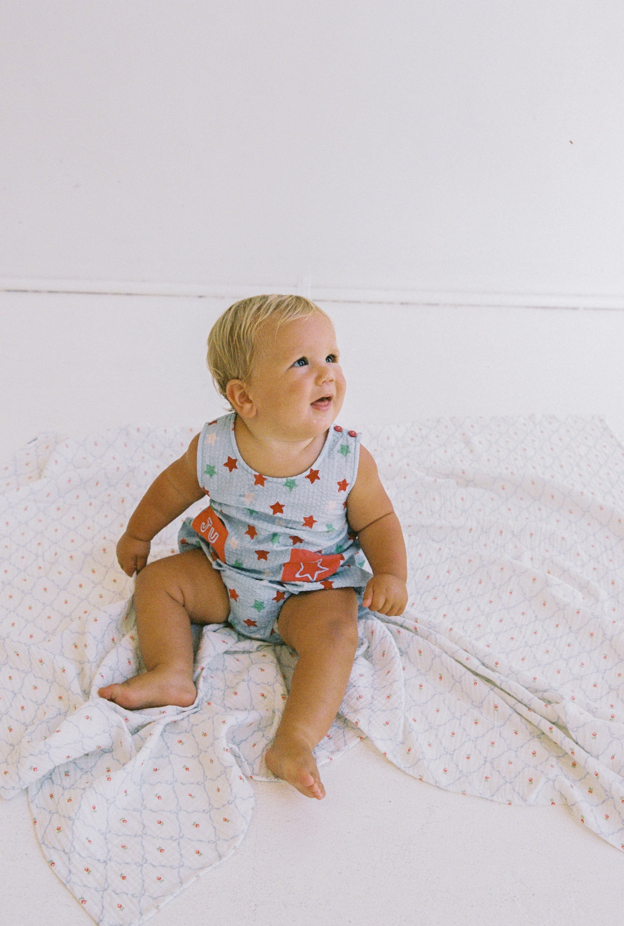 A baby with blonde hair sits on JUNI JNR's Levi Romper, a soft, patterned blanket. Clad in the Levi Romper—a striped outfit featuring red stars, a crab design, and functional pockets—the baby gazes upward joyfully against a simple white wall background.