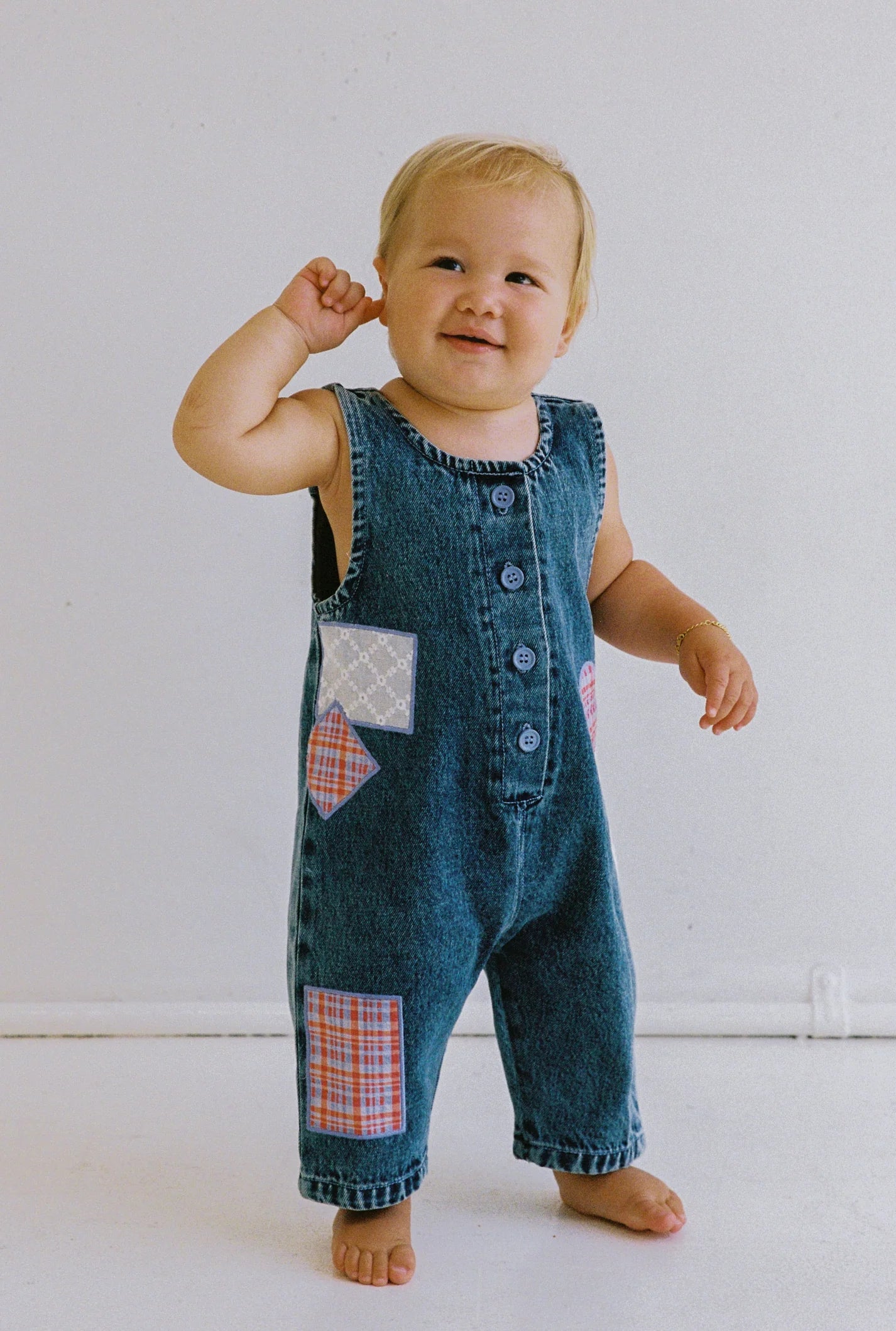 A smiling toddler stands on a white backdrop in JUNI JNR's Charlie Overalls, an ethically made denim jumpsuit with vibrant patchwork designs. The child, barefoot with light blonde hair and one hand near the ear, adds charm to the delightful ensemble.