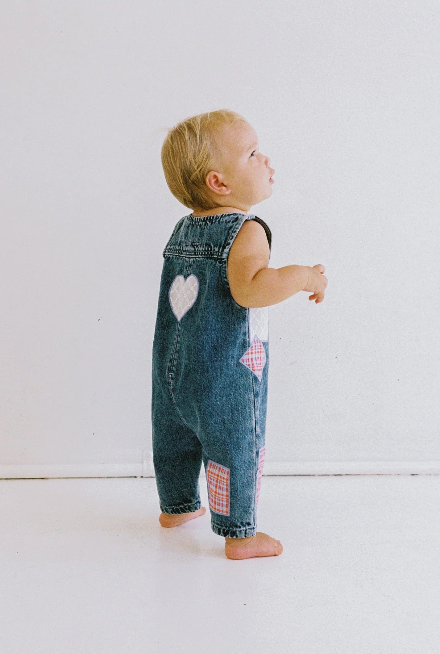 A blonde toddler stands barefoot against a white background, wearing JUNI JNR ~ Charlie Overalls by JUNI JNR. The ethically made denim features heart and patchwork designs with functional buttons, adding charm as the child looks upwards, slightly turned to the side.