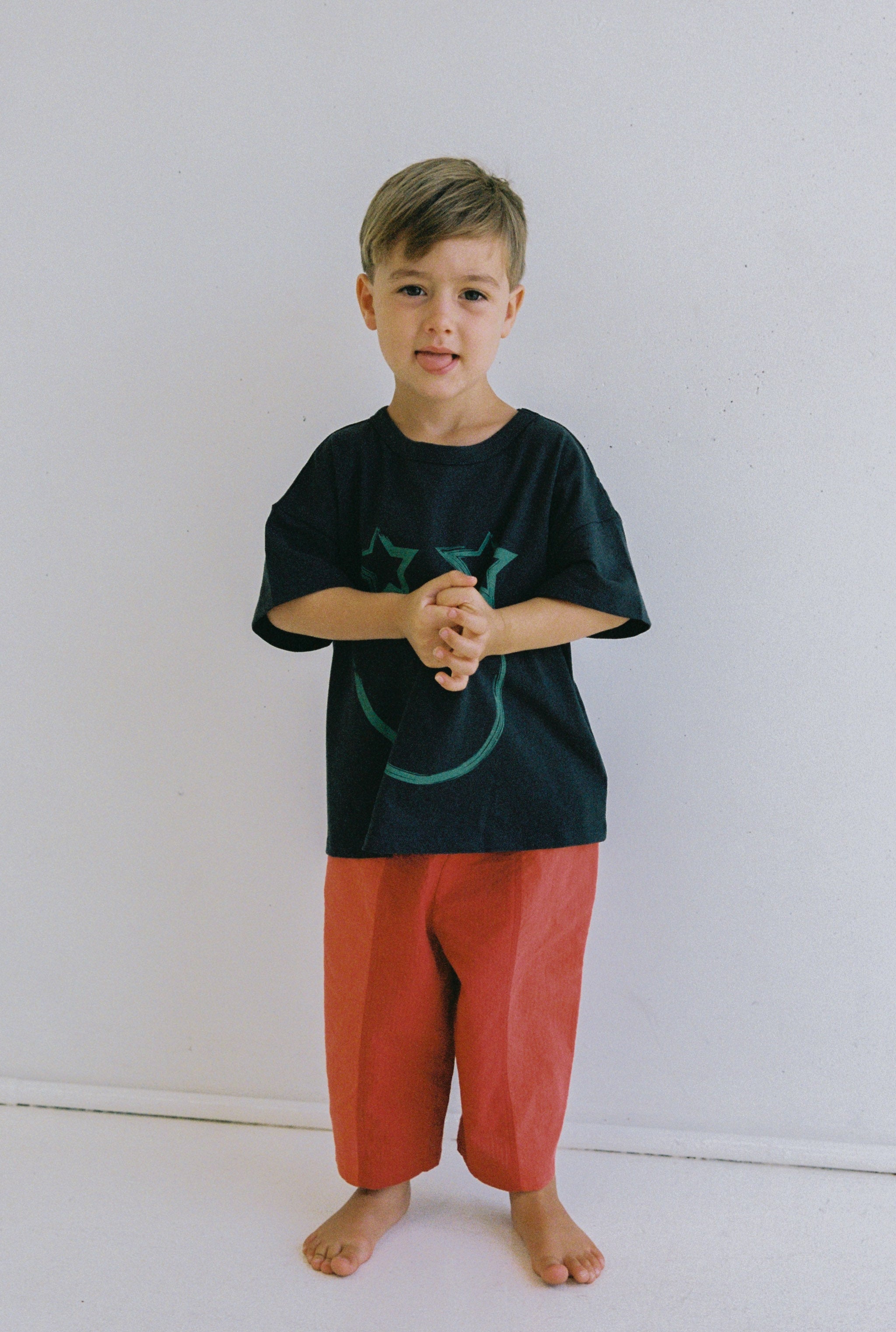 A young child stands barefoot against a plain white background, wearing a loose black t-shirt with a subtle design and the vibrant Otis Pant in bright red featuring an elastic waistband. The ensemble from JUNI JNR reflects an embrace of comfort and style, capturing the essence of ethically made fashion.