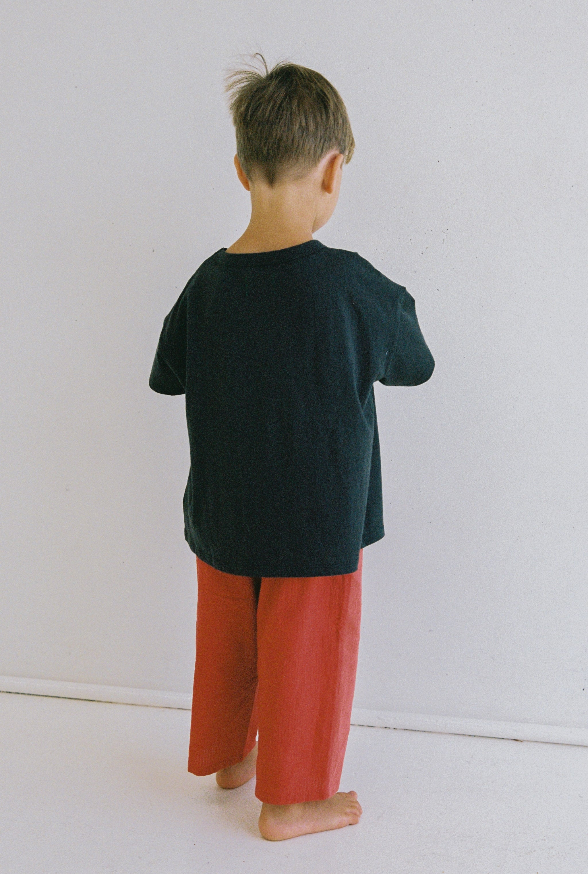 A young child stands barefoot against a plain white wall, facing away. They wear the Stellar Tee by JUNI JNR—a dark blue shirt—and loose red pants crafted from ethically made, organic cotton. The child's hair is short and light brown.