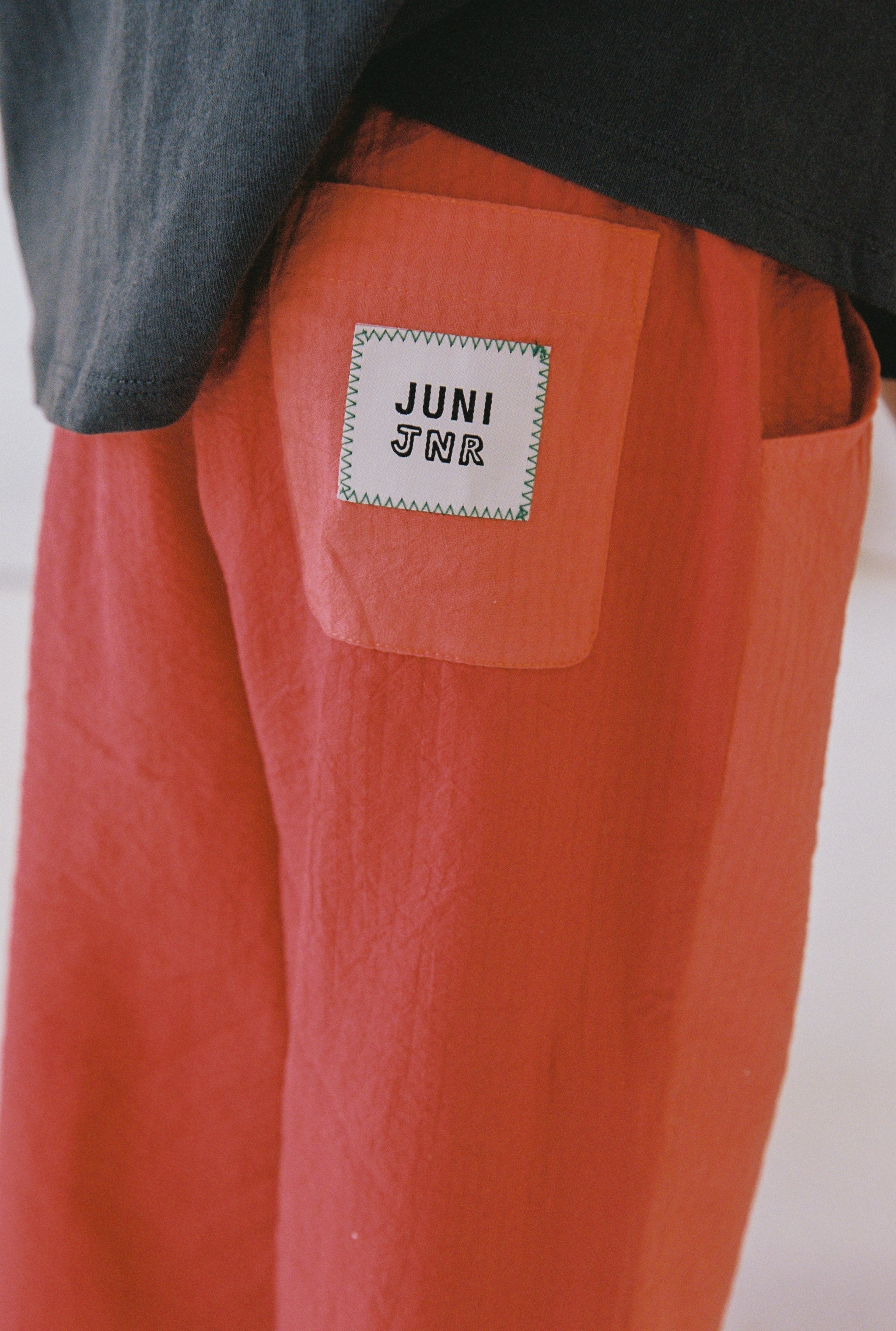 Close-up of a person wearing the Otis Pant, styled in red from ethically made cotton seersucker fabric, featuring a pocket on the right side. A white patch on the pocket proudly displays "JUNI JNR" in bold text. The person is also wearing a black top, which is partially visible in the frame.