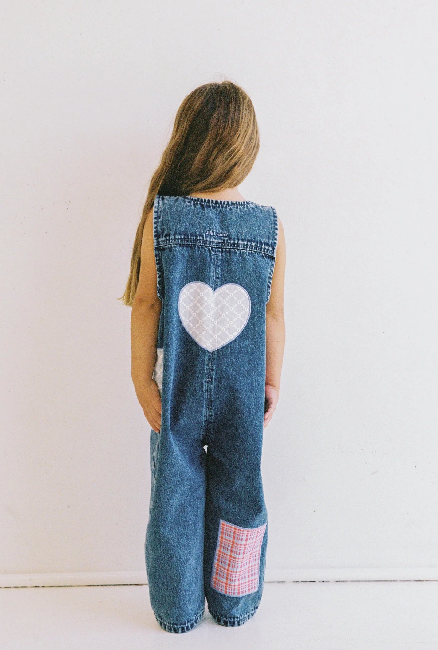 A young child with long hair, facing away against a plain white backdrop, wears ethically made JUNI JNR Charlie Overalls in blue denim. These overalls stylishly feature a white heart patch on the back and a red checkered patchwork near the leg, blending style and sustainability.