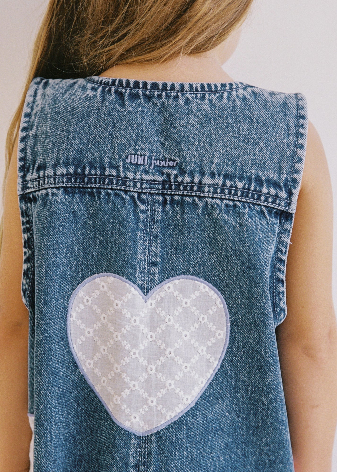 A child wears JUNI JNR ~ Charlie Overalls, featuring a unique sleeveless denim design with a large white heart patch on the back and the brand "JUNI JNR" proudly displayed above, celebrating ethical patchwork artistry.