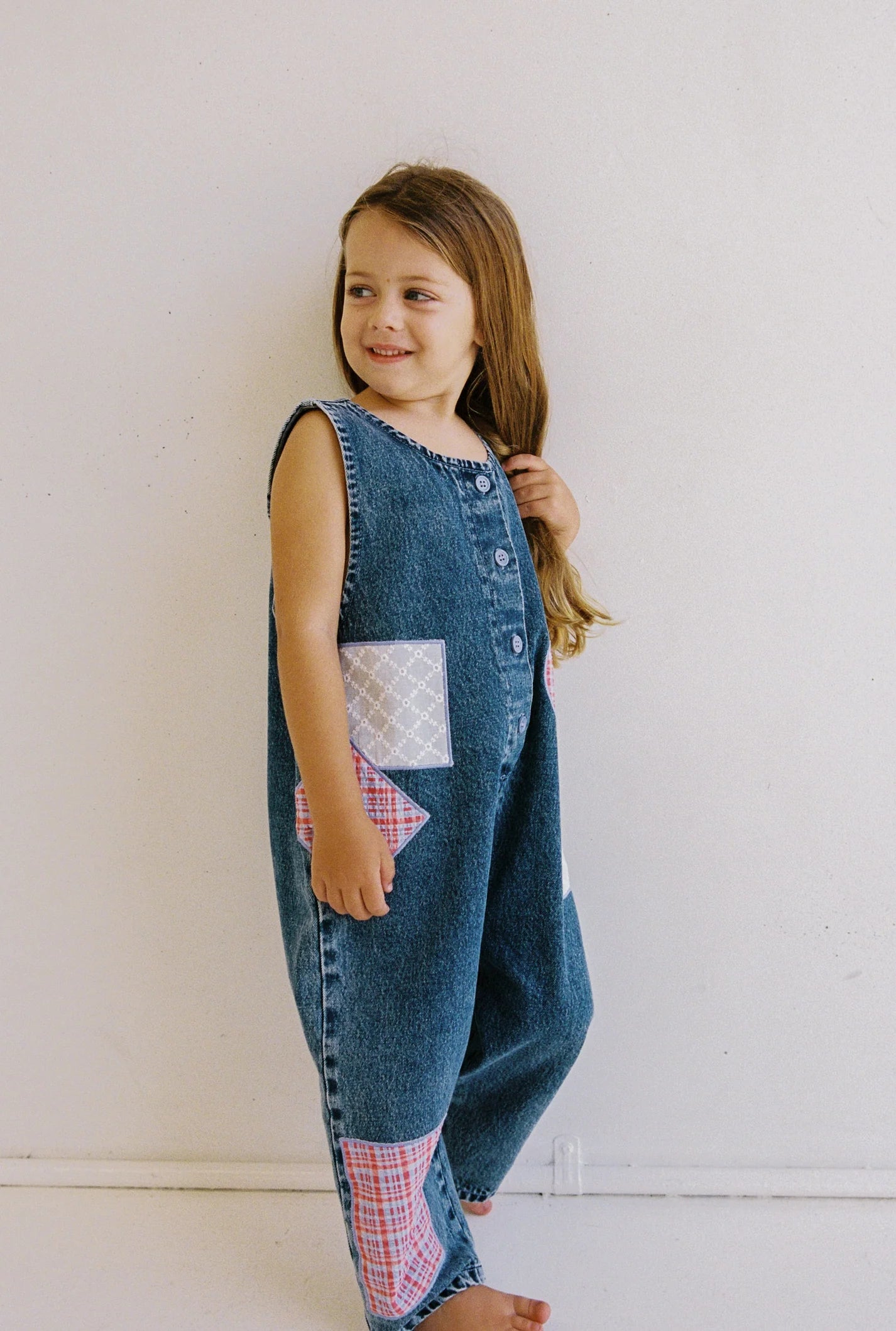 A young girl with long hair stands barefoot against a light wall, smiling and looking to the side. Her Charlie Overalls by JUNI JNR showcase functional buttons, adding practicality to its charming patchwork denim design.