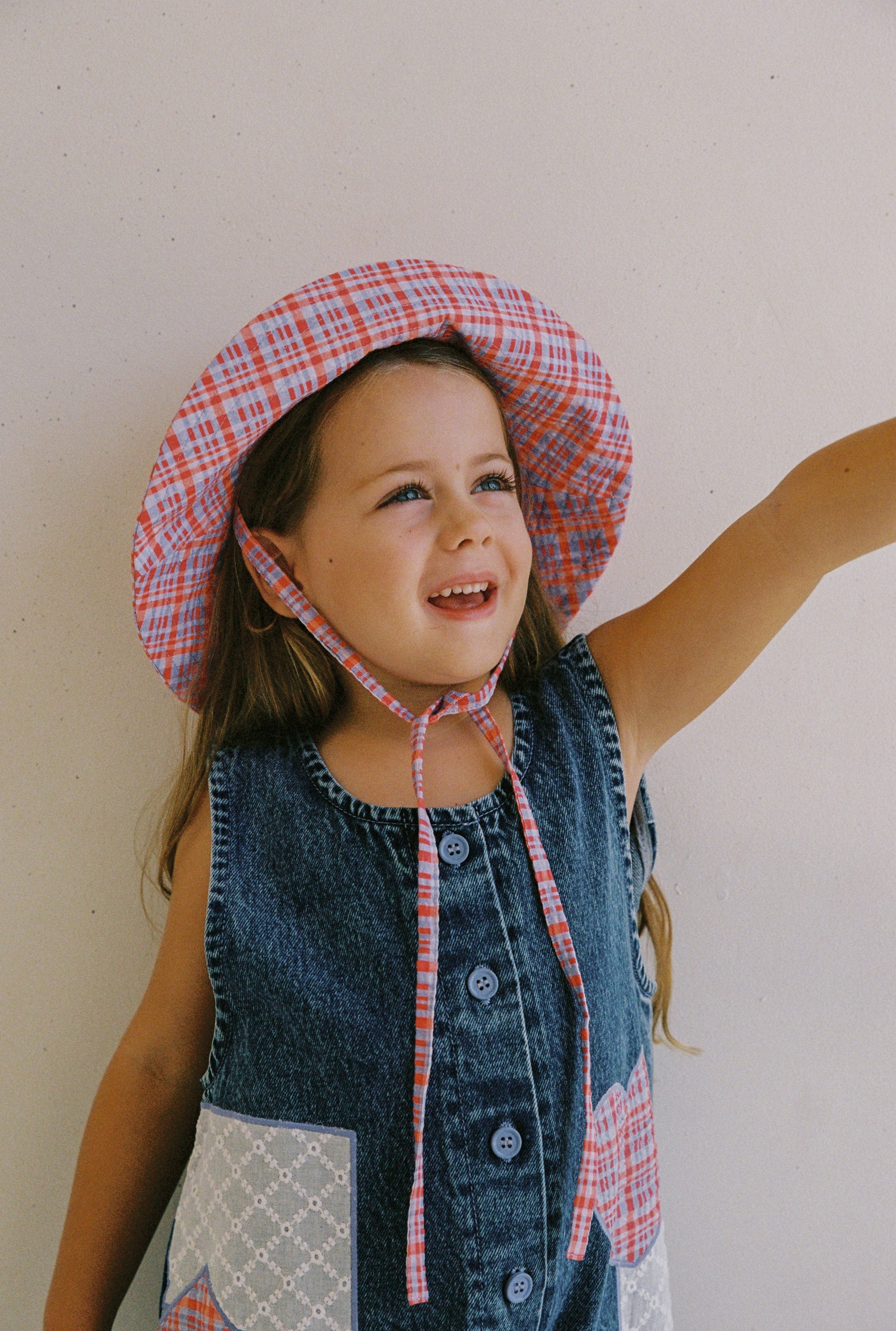 A young girl in a denim dress with buttons and the JUNI JNR Picnic Check Sunhat smiles and points upwards. Made from ethically sourced materials, the dress has white patterned pockets. She stands against a light-colored wall, appearing cheerful and curious.