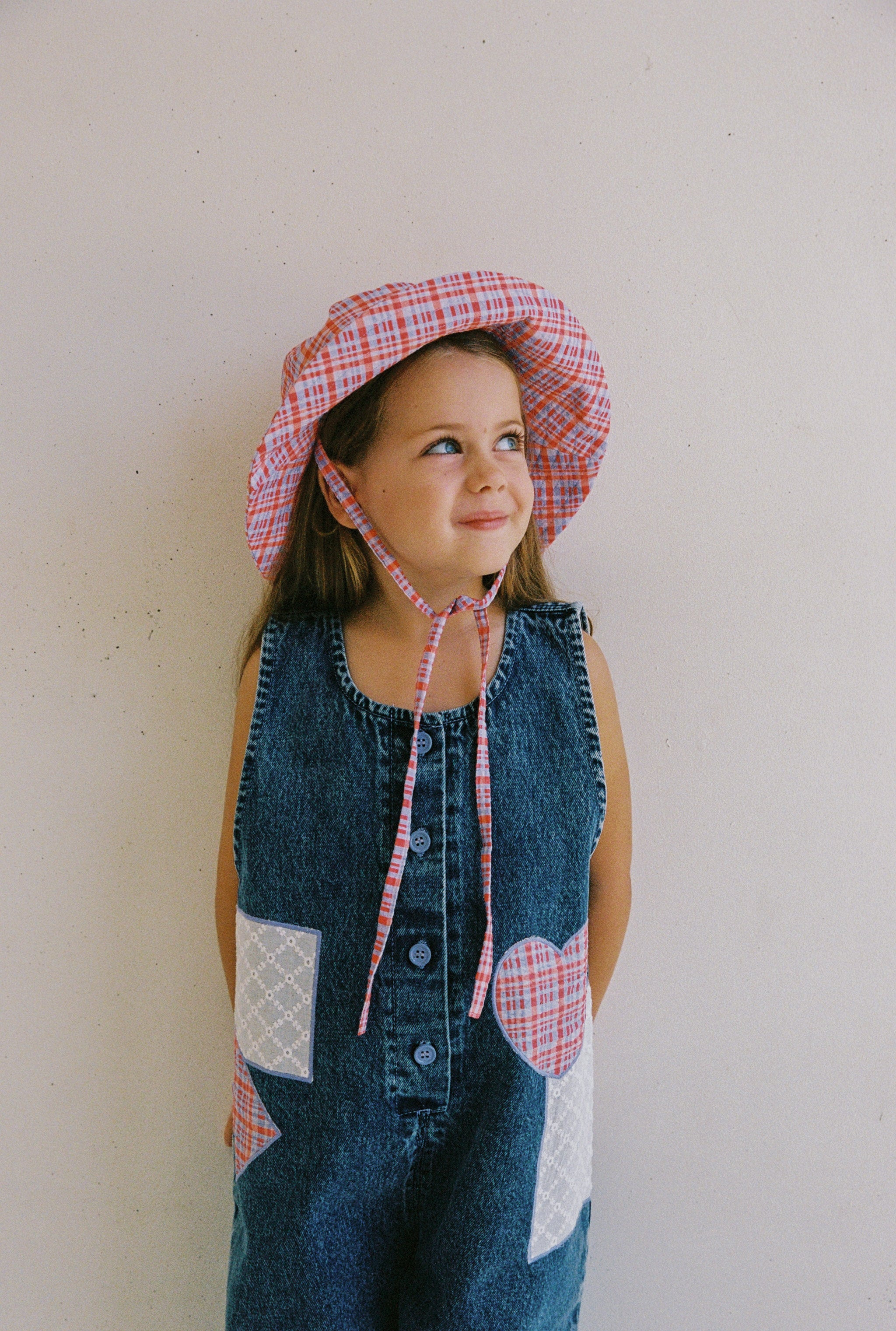 A young girl poses thoughtfully against a plain, light-colored wall, wearing a denim dress with pink and white checkered heart patches and the matching "Picnic Check Sunhat" by JUNI JNR. Her outfit, ethically made in a BSCI-certified factory, adds extra charm to her appearance.