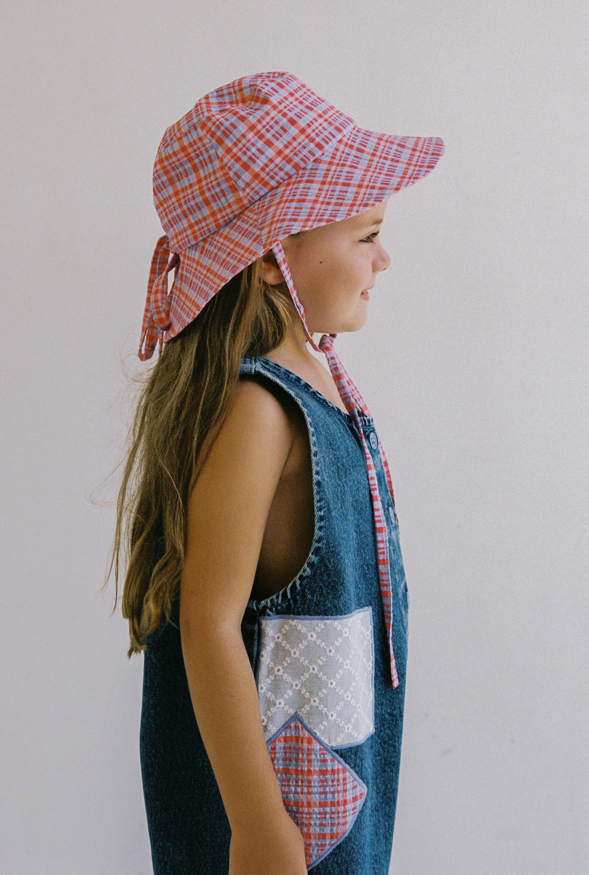 A girl with long hair wears a JUNI JNR Picnic Check Sunhat and a blue sleeveless denim dress made from seersucker fabric, featuring two diamond-pattern pockets. She stands sideways against a plain background, smiling, and showcases an ethically made style.