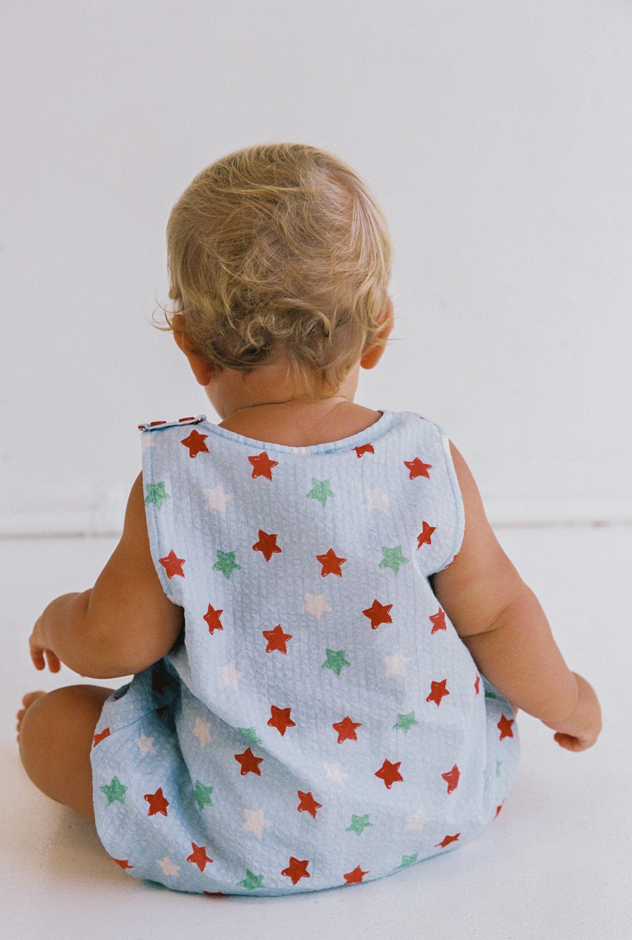 A baby with curly blonde hair is sitting on the floor, facing away. The Levi Romper from JUNI JNR stands out in blue with red, white, and green stars and is made from soft seersucker fabric. The background features a plain white wall.