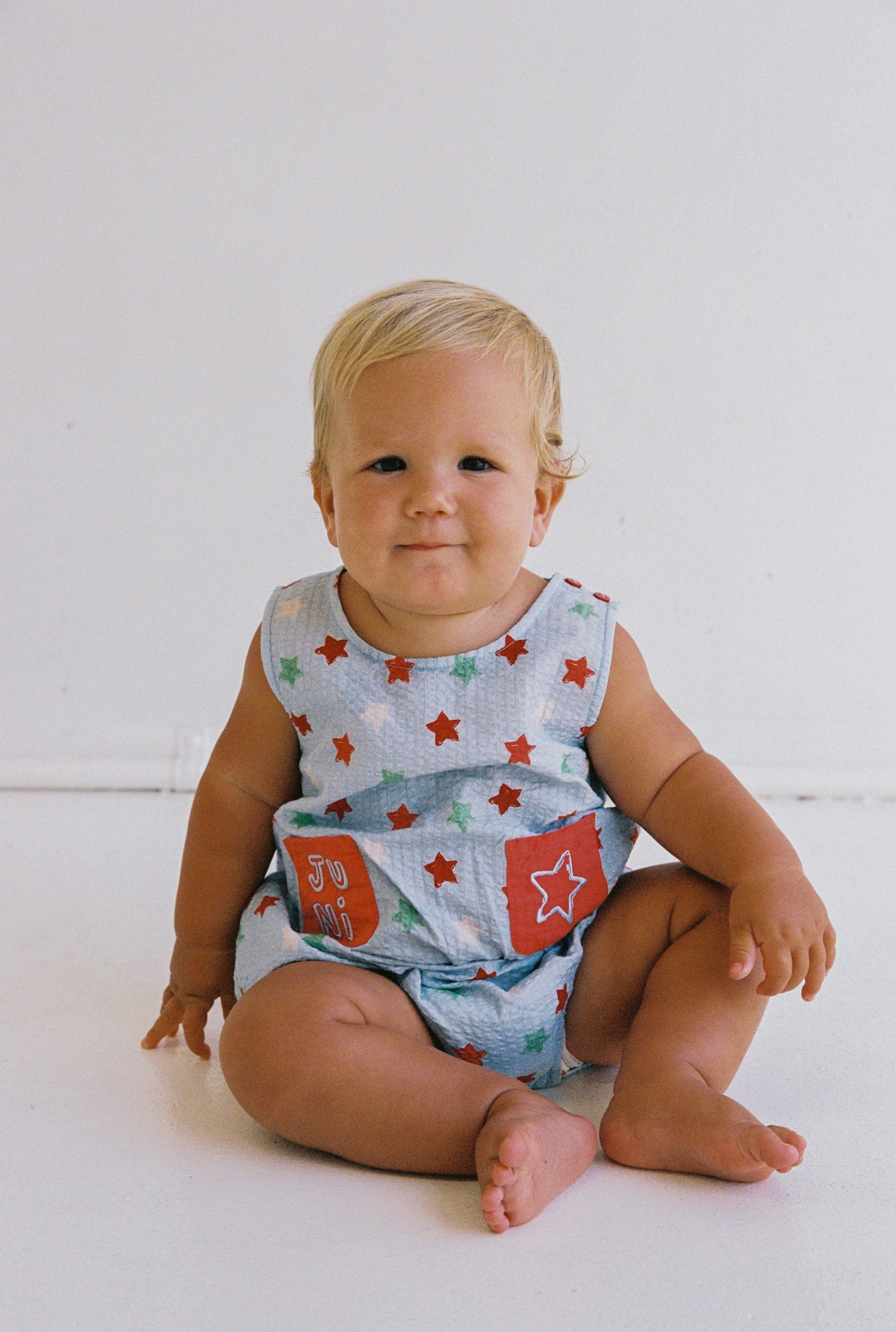 A smiling baby with blond hair sits on the floor wearing the Levi Romper by JUNI JNR, featuring a blue outfit adorned with red stars, patches, and functional pockets. The ethically made seersucker fabric ensemble stands out against a plain white wall.