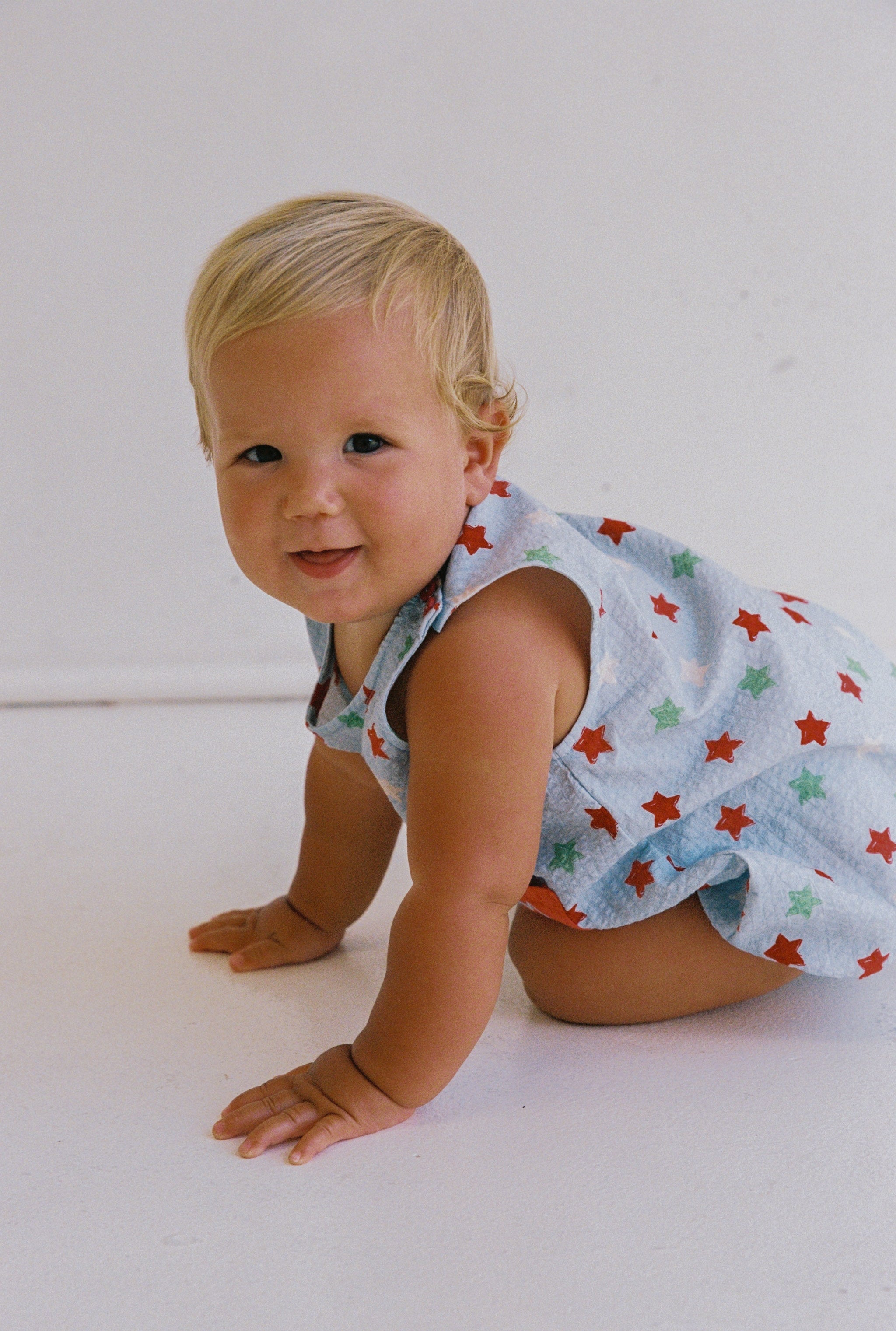 A baby with blonde hair, wearing a Levi Romper by JUNI JNR in light blue with red and green stars, is crawling on a white seersucker fabric surface while looking at the camera.