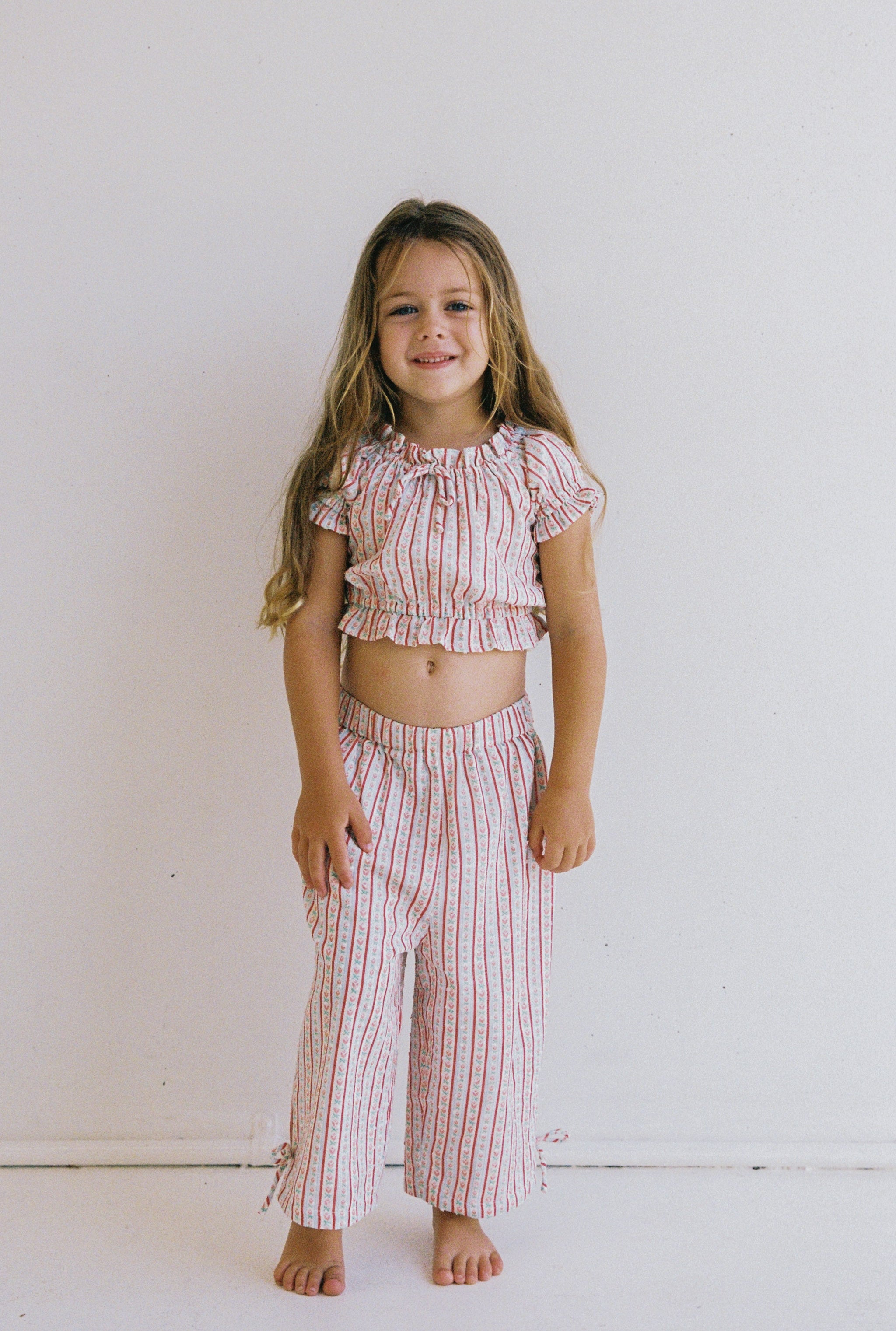 A young girl with long hair smiles while standing against a plain white background. She is wearing the Dahlia Top by JUNI JNR, a matching outfit made of textured cotton fabric, featuring a short-sleeve ruffled top and wide-leg pants with a red and white striped pattern.