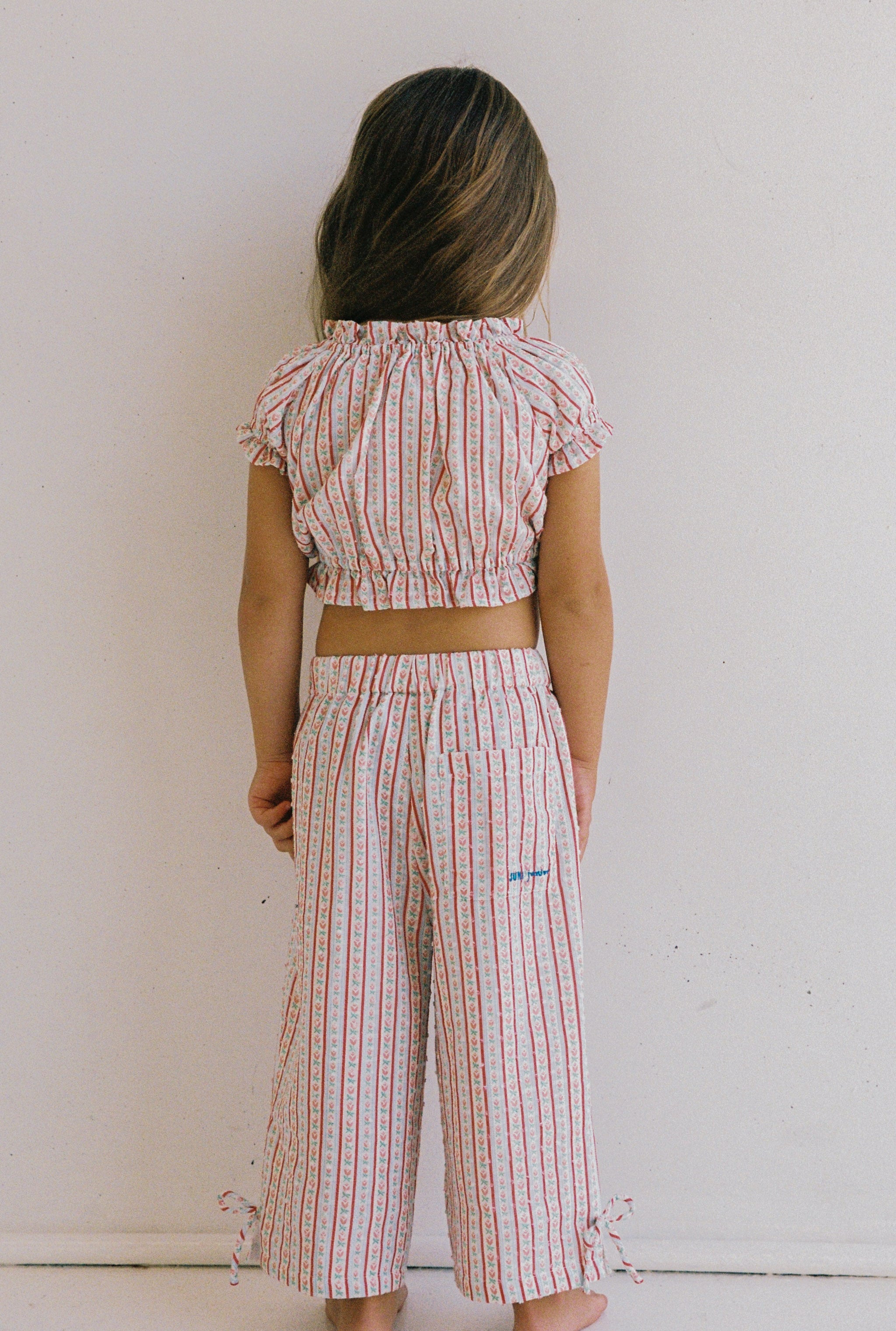 A child with long hair stands facing away, dressed in a matching outfit made from textured cotton fabric with a striped pattern. The Dahlia Top by JUNI JNR is cropped with short sleeves, and the loose-fitting pants include ties at the hem. This ethically crafted ensemble stands out beautifully against the plain white wall background.