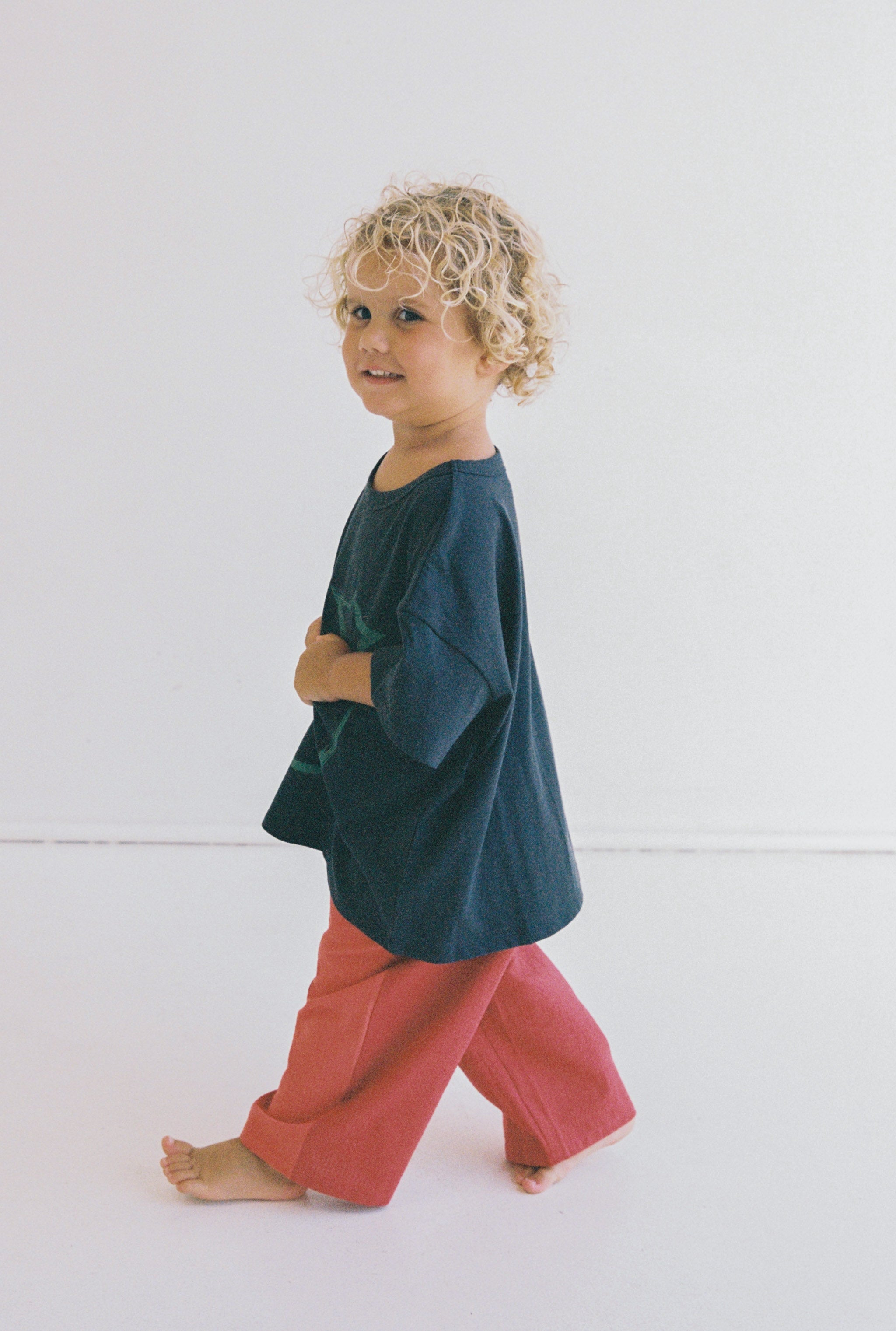 A young child with curly blond hair is joyfully walking barefoot, wearing an oversized Stellar Tee by JUNI JNR. The shirt, made from ethically produced organic cotton, features green designs on a navy blue background and is paired with red pants. The plain white backdrop accentuates the child's playful movement.