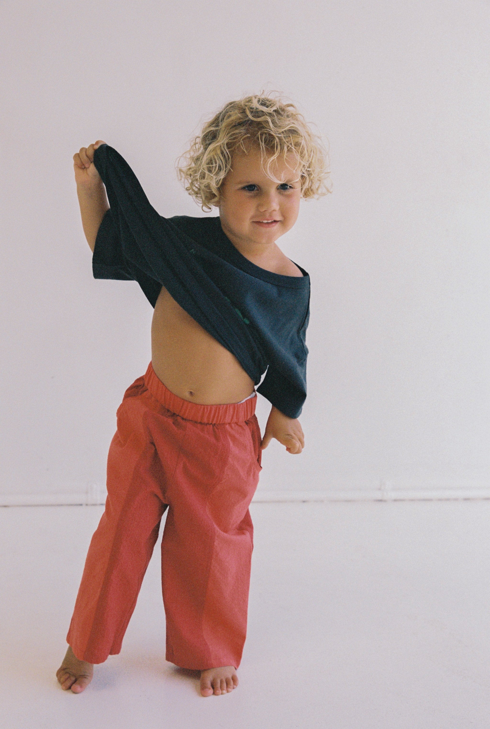 A child with curly blonde hair playfully lifts their black shirt, revealing their belly. They are wearing the Otis Pant by JUNI JNR, featuring an elastic waistband in a striking red color, and are barefoot, standing against a plain white background.