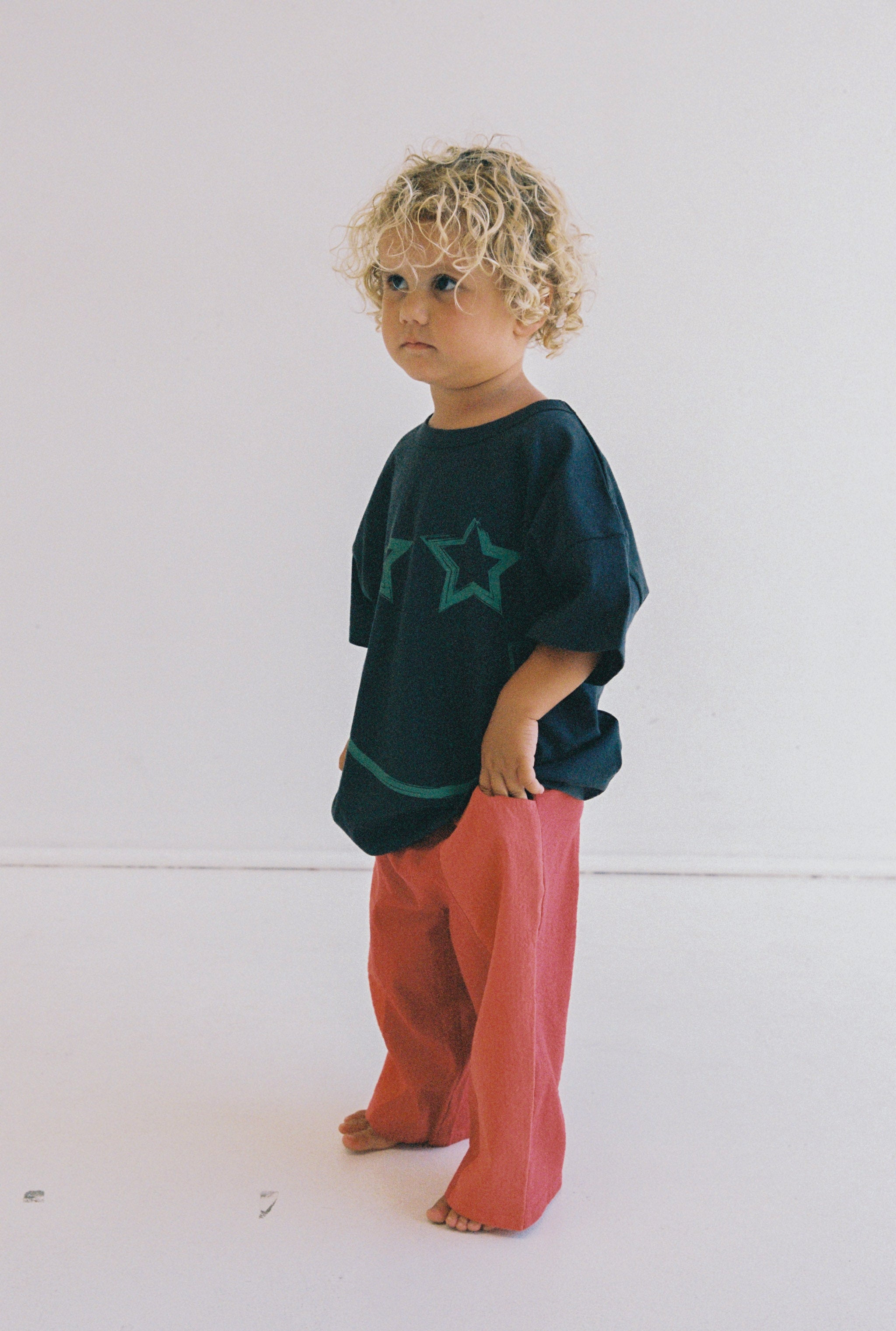 A young child with curly hair stands barefoot against a plain background, wearing the Stellar Tee from JUNI JNR. The black shirt is decorated with star designs and paired with red pants. This outfit is crafted from GOTS certified organic cotton, ensuring it is ethically made for both style and comfort.