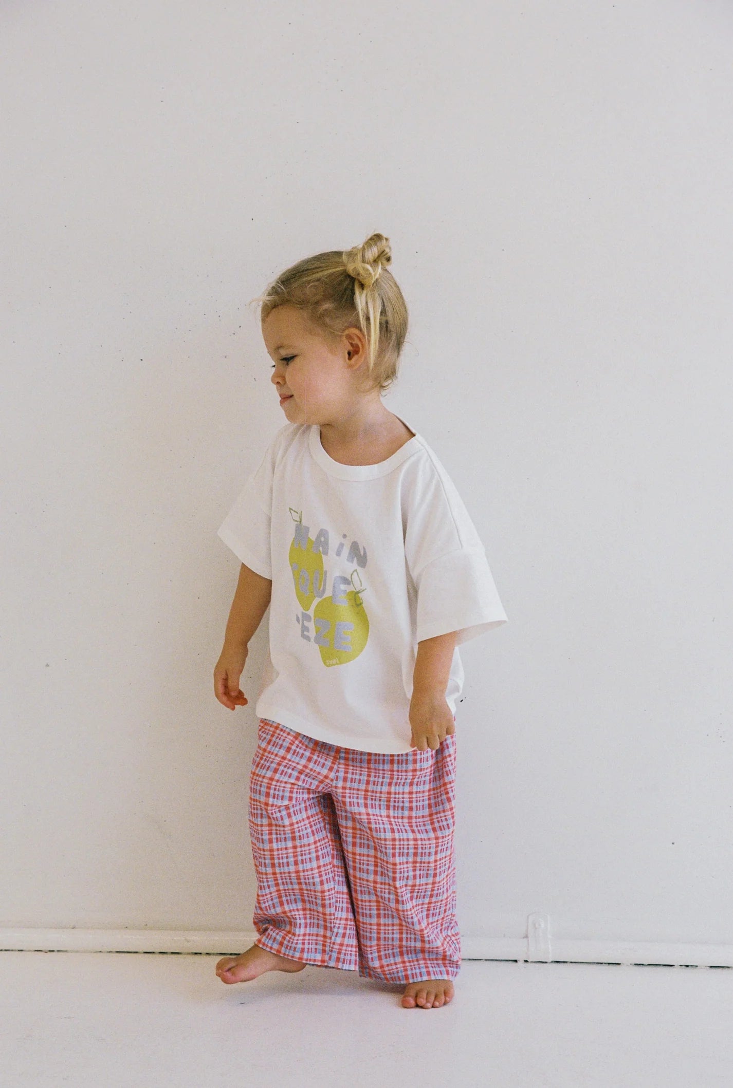 A young child with blonde hair in a bun stands against a white wall, wearing a white T-shirt with a yellow graphic and text. They are dressed in JUNI JNR's ethically made Lenny Pant Picnic Check, featuring a red and blue plaid design with an elastic waistband. The child gazes downward and slightly to the side.