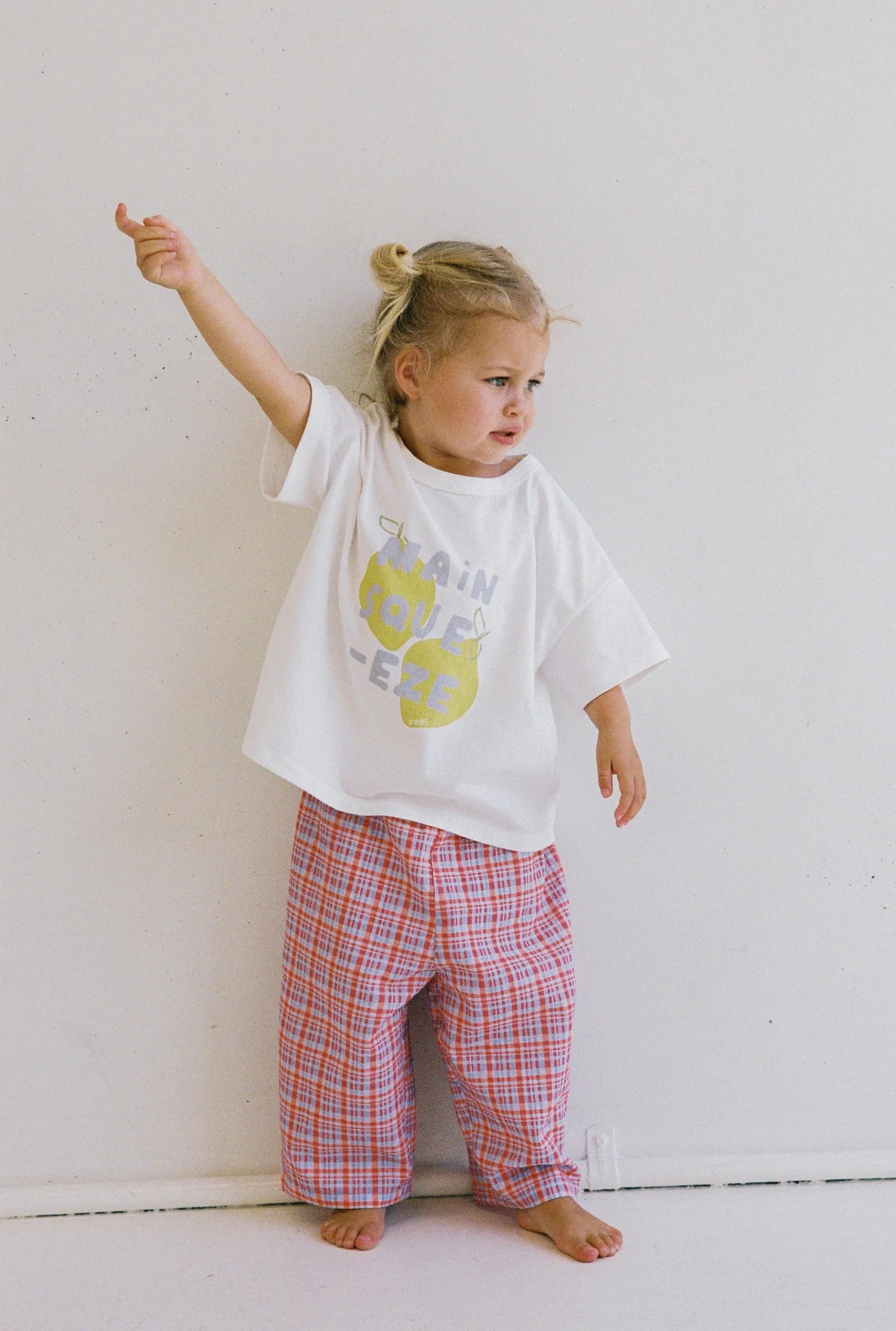 A young child stands against a plain wall, wearing an ethically made white T-shirt with abstract graphics and JUNI JNR's Lenny Pant Picnic Check. The oversized pink plaid pants feature an elastic waistband. The child raises their arm, looking to the side thoughtfully.