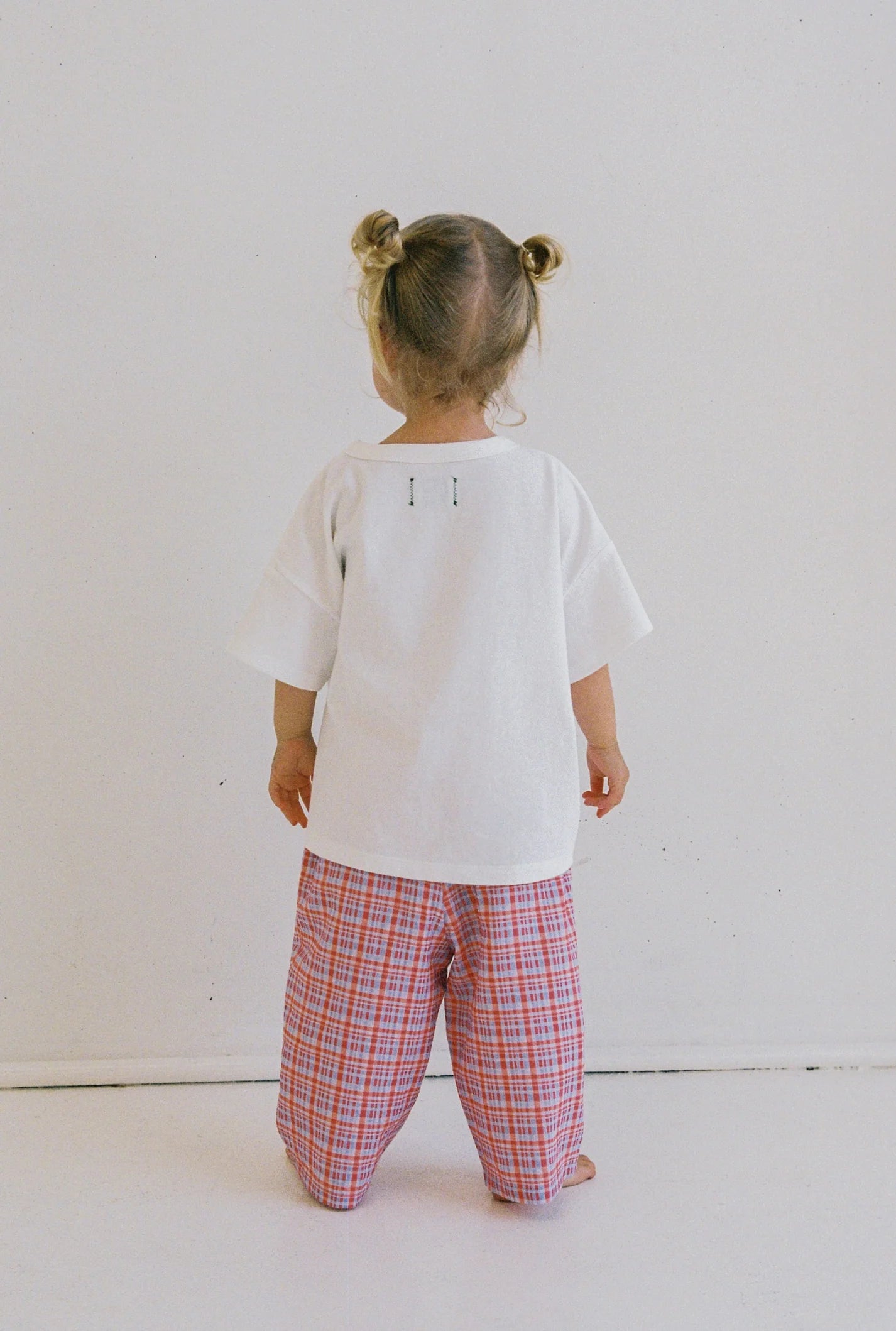 A toddler stands with their back to the camera, wearing JUNI JNR's Lenny Pant Picnic Check, featuring an elastic waistband and pink plaid design. Their hair is styled in small buns against a plain off-white wall and floor, enhancing the ethically made outfit from JUNI JNR.