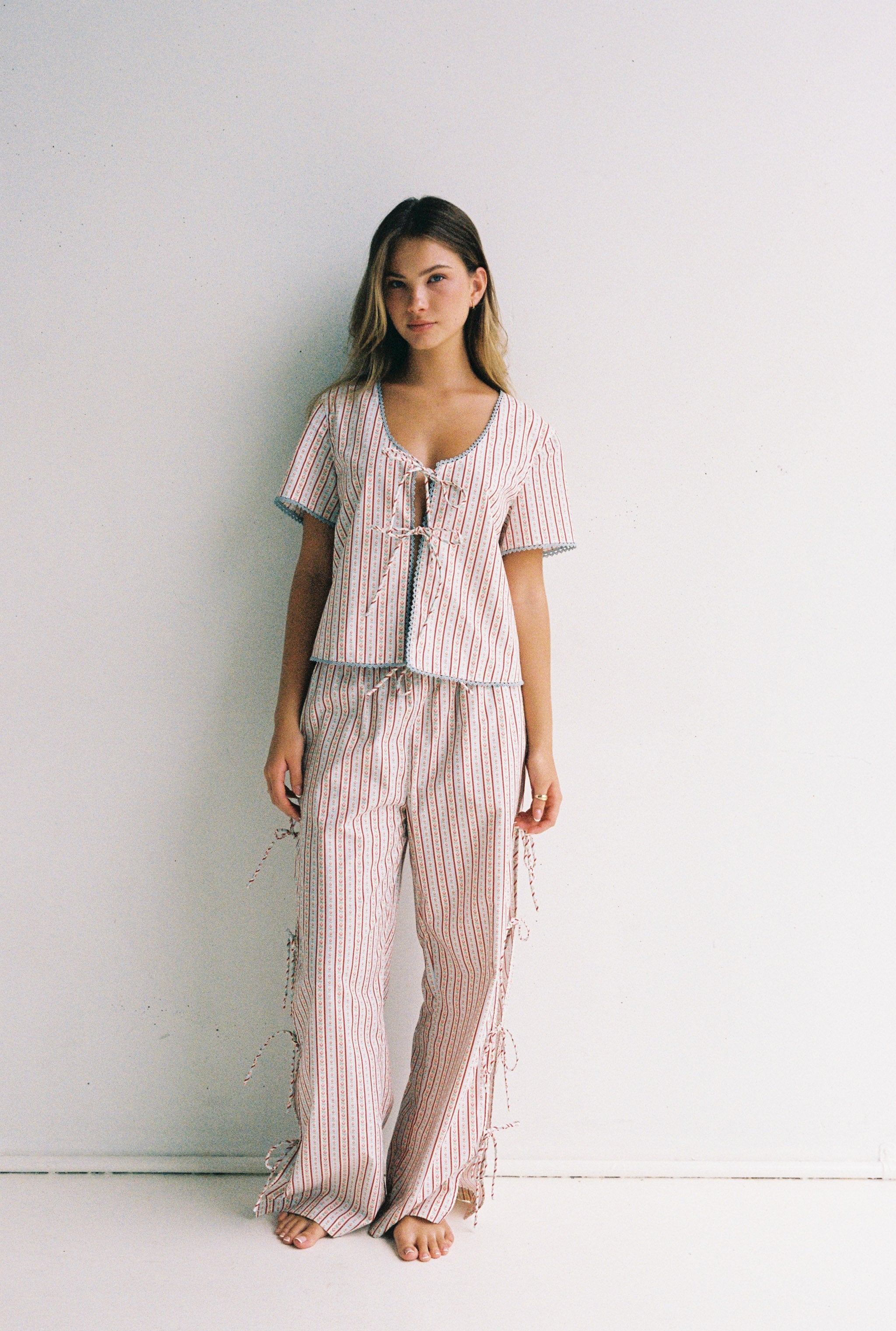 A person stands barefoot against a plain white background, wearing the Poppy Blouse from JUNI, a matching two-piece outfit with vertical stripes. This cotton poplin ensemble features a scooped neckline and includes a short-sleeved top paired with pants. They are looking directly at the camera.